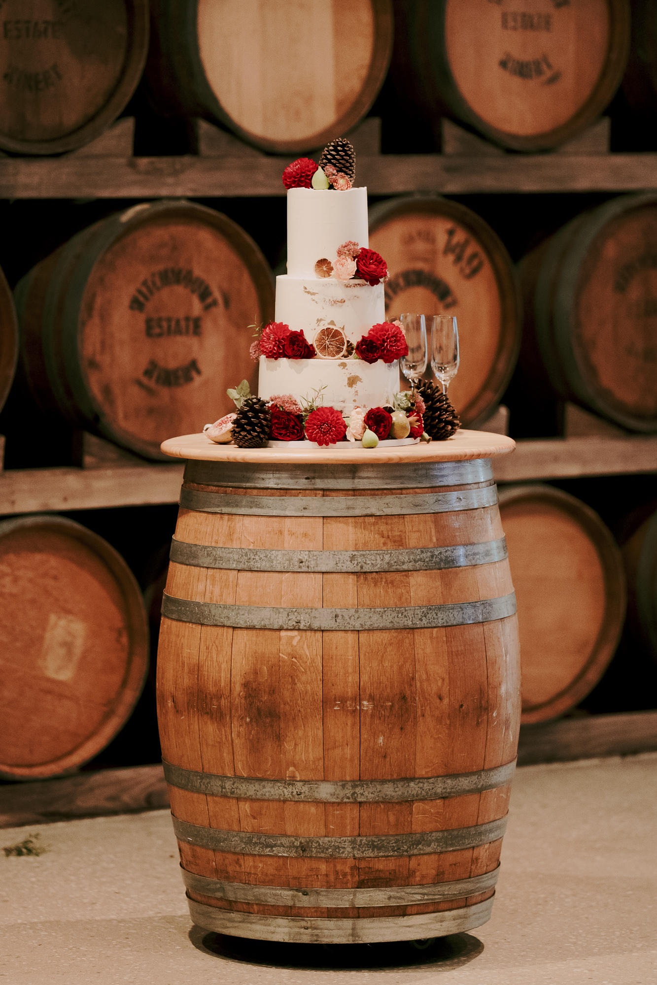 A three-tiered white wedding cake decorated with red and brown flowers, pinecones, and dried citrus slices, sitting on top of a wooden barrel in a wine cellar or winery with large barrels in the background. Witchmount Estate