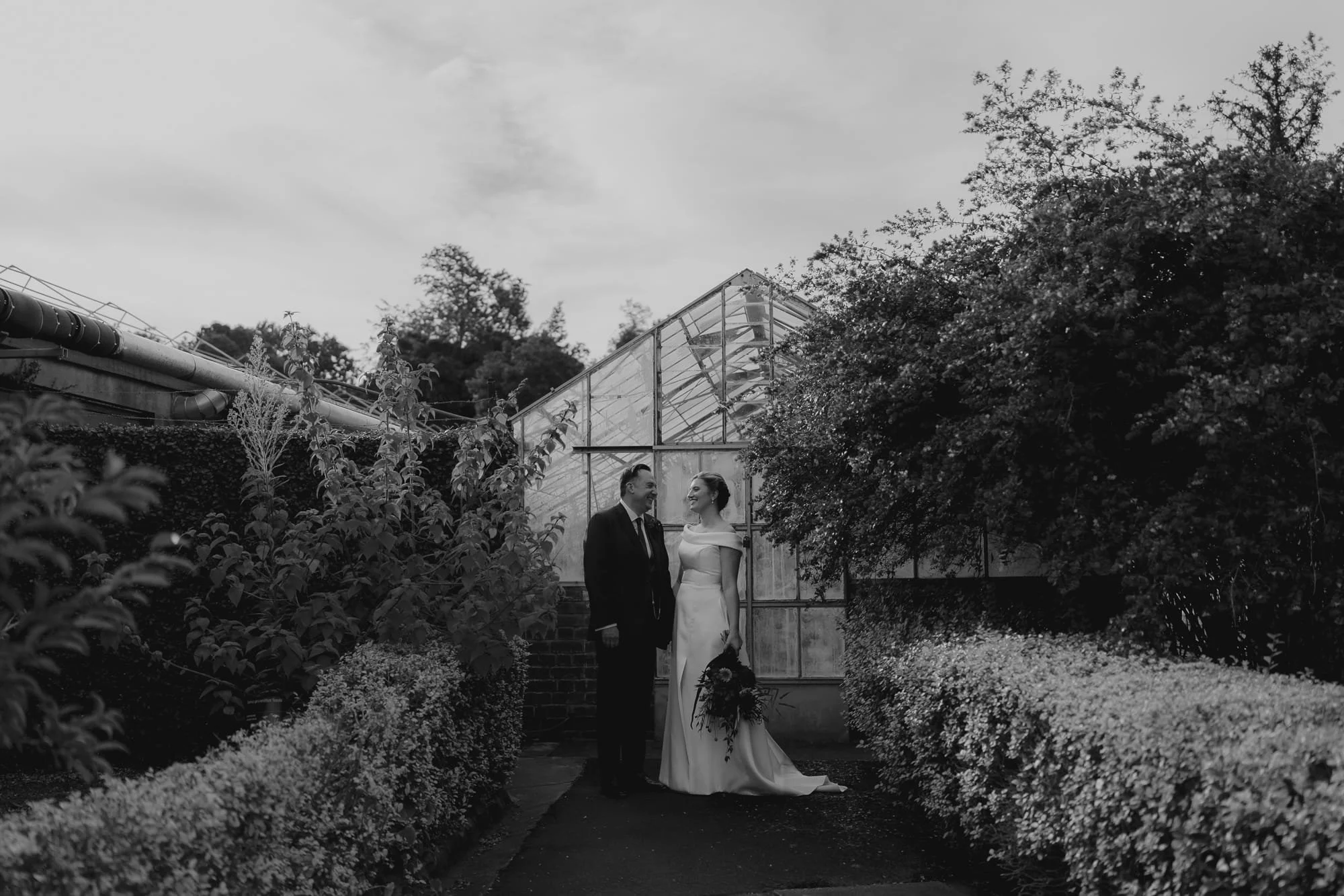 Melbourne Wedding Photography. Black and white photo of a bride and groom standing together in the Royal Botanical Gardens Melbourne with a greenhouse in the background. The bride is holding a bouquet and is dressed in an off-the-shoulder gown. 