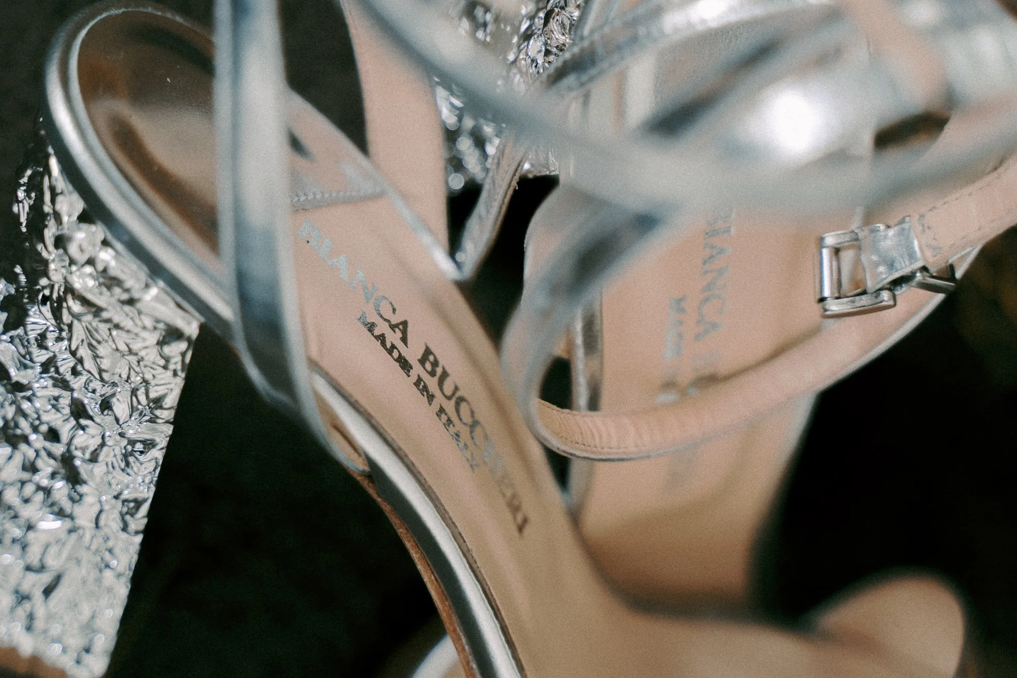 Close-up of a pair of silver high-heeled shoes with beige insoles, placed next to a shiny, textured silver clutch purse.