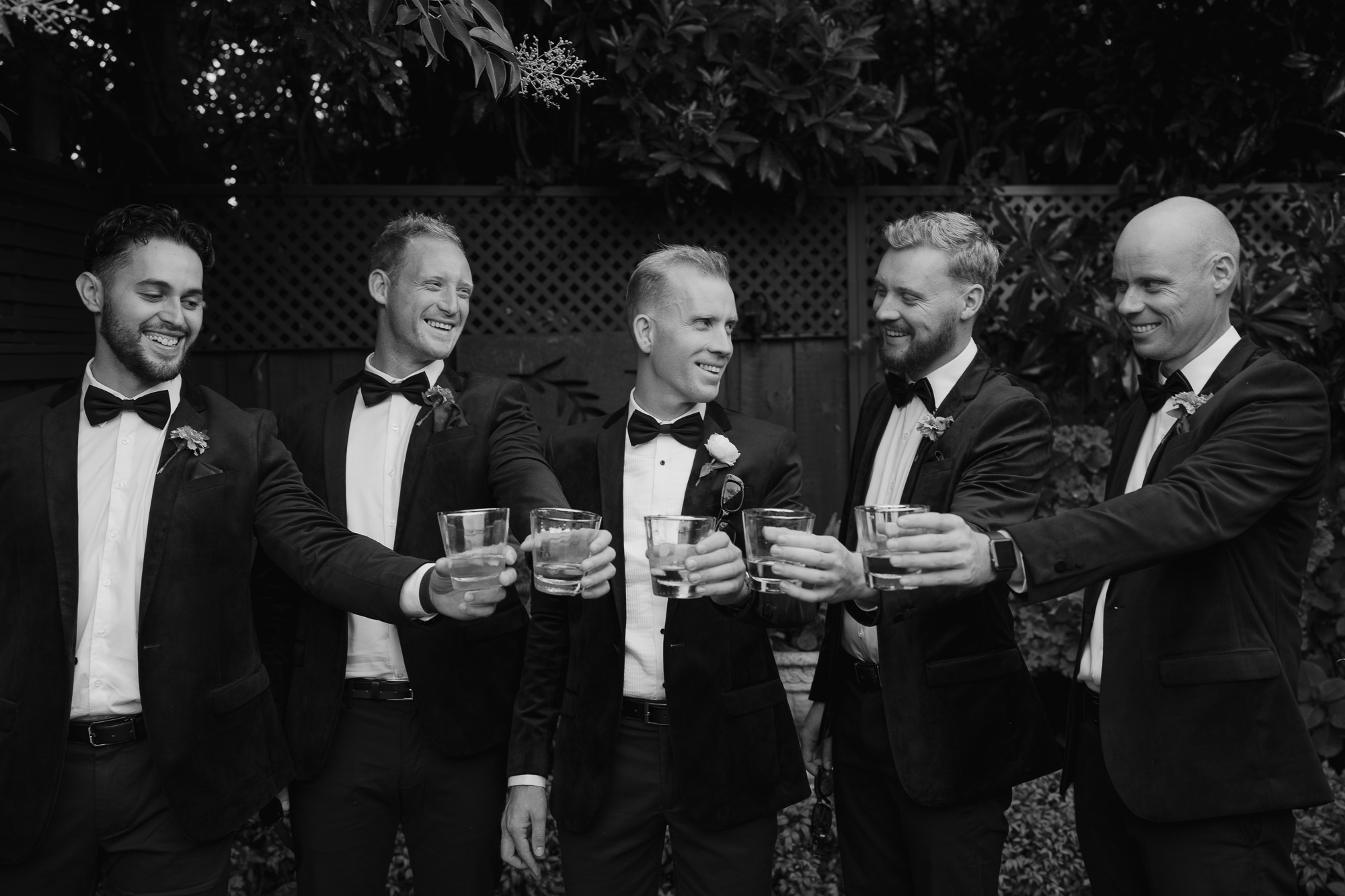 A group of six men dressed in tuxedos and bow ties celebrating with drinks at an outdoor gathering.