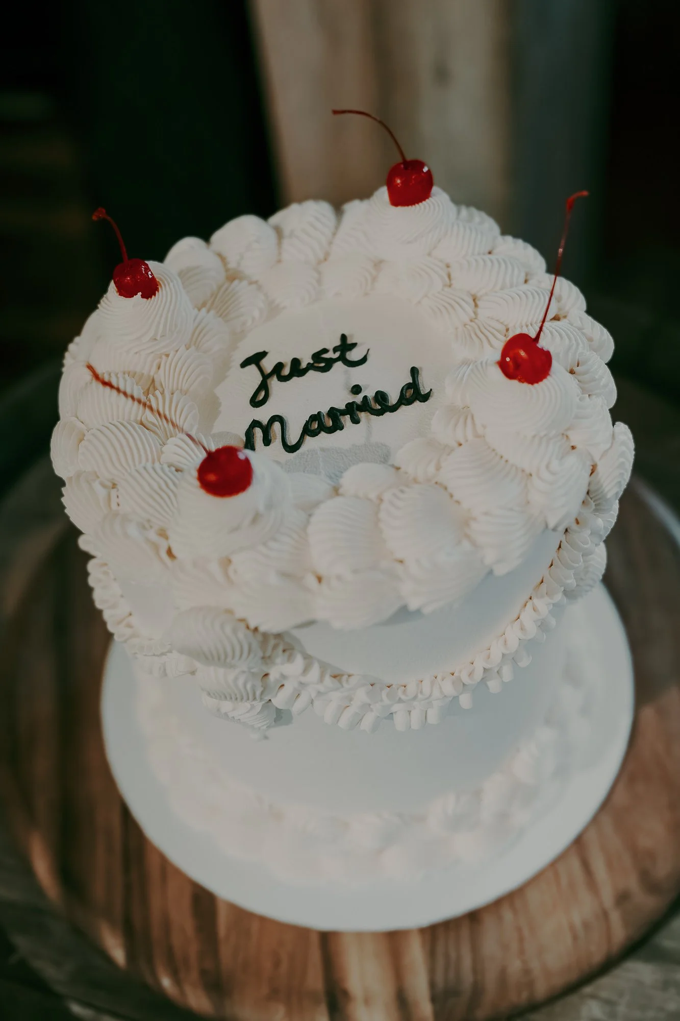 A white wedding cake with piped frosting, topped with cherries, and inscribed with "Just Married" in black icing.