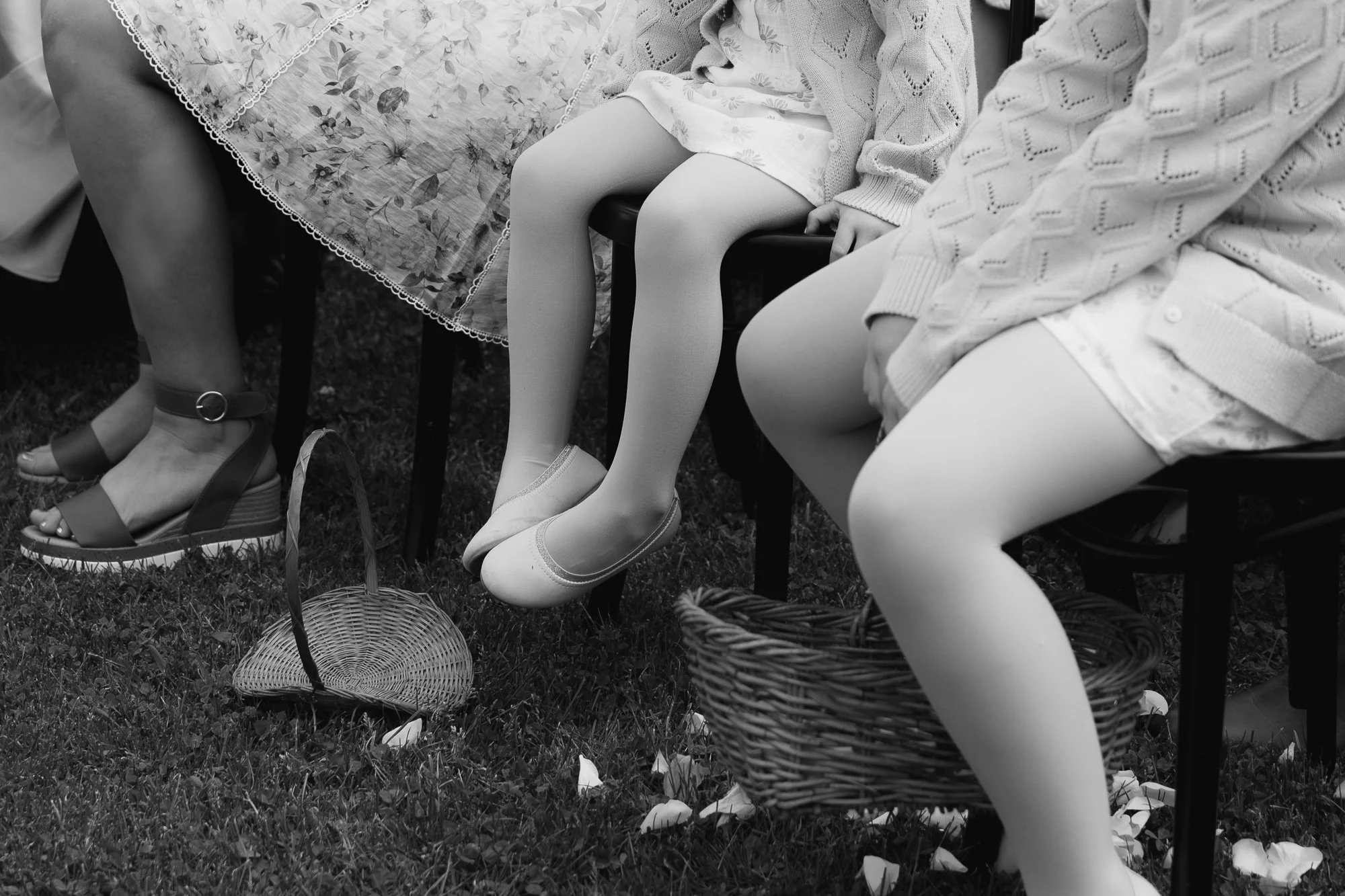 People sitting on benches with their legs dangling, wearing dresses and tights, with empty baskets on the ground, during a wedding ceremony at the Vines on Avon.