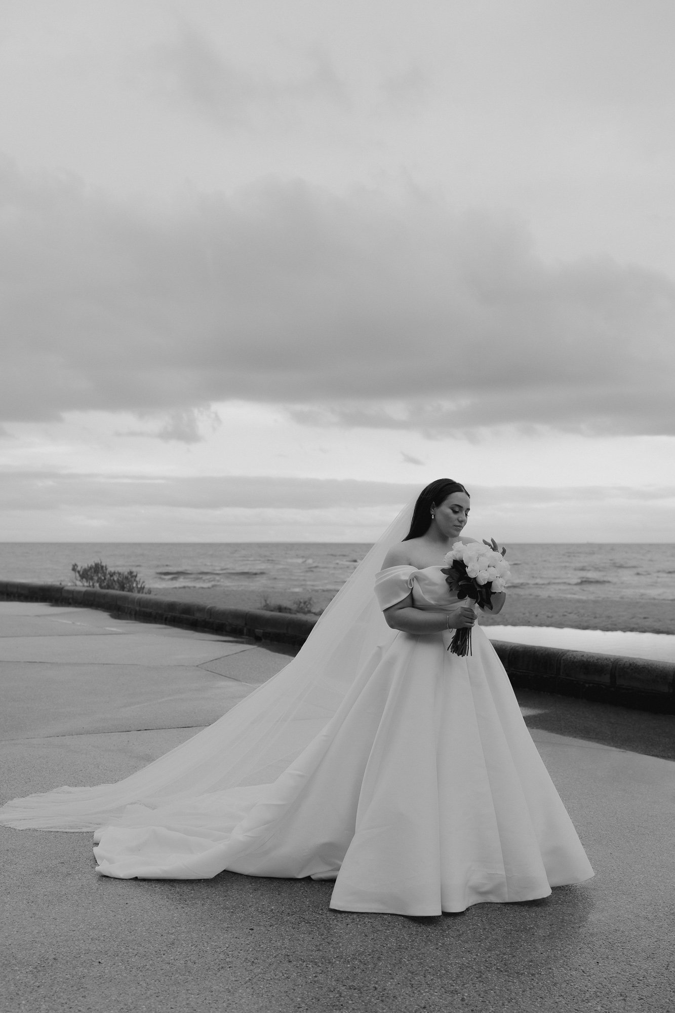 A bride in a wedding dress holding a bouquet, standing outdoors near the ocean on a cloudy day.