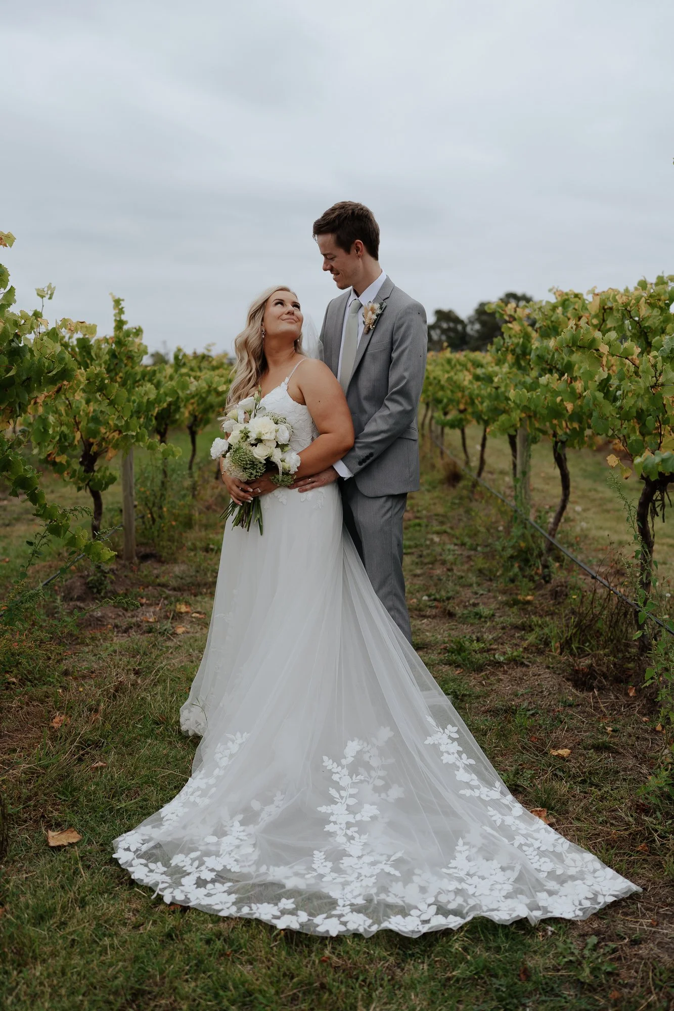 Bride and groom standing in a vineyard at the Vines of Avon, Stratford, under cloudy skies, gazing at each other. The bride wears a white wedding gown with a long train and holds a bouquet of white flowers. The groom is dressed in a gray suit.