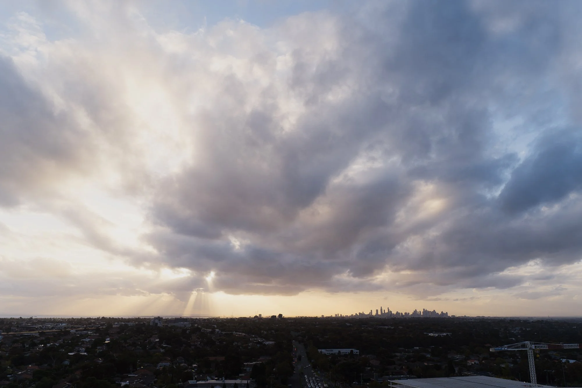 A city skyline under a cloudy sky, with sunlight breaking through the clouds. Taken from Altus Event Space at the Chadstone Hotel.