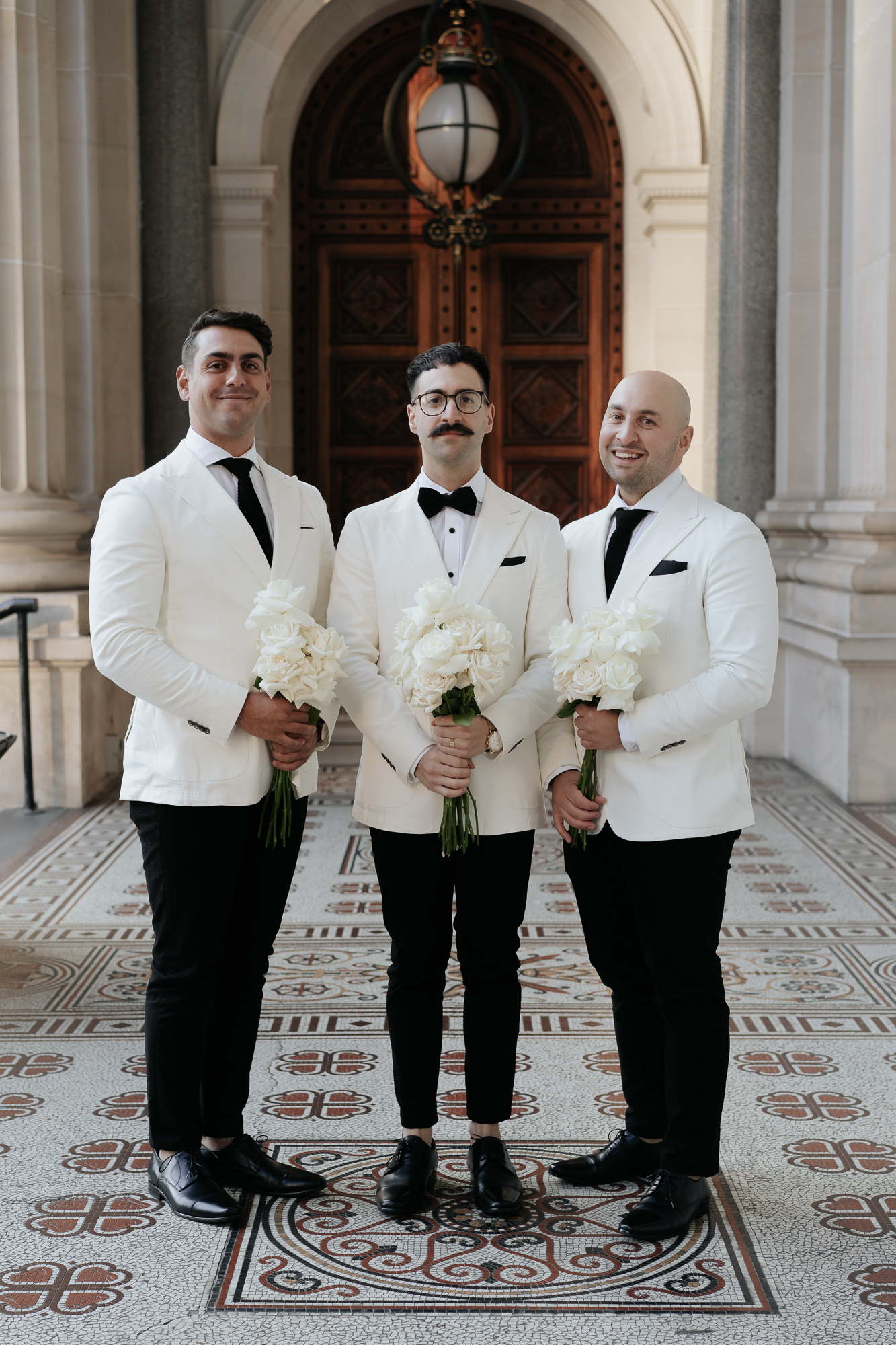 Three men dressed in white tuxedo jackets, black pants, and black shoes, holding white rose bouquets, standing in front of a grand arched wooden doorway inside a historic building with ornate mosaic tile flooring. Parliament House Melbourne