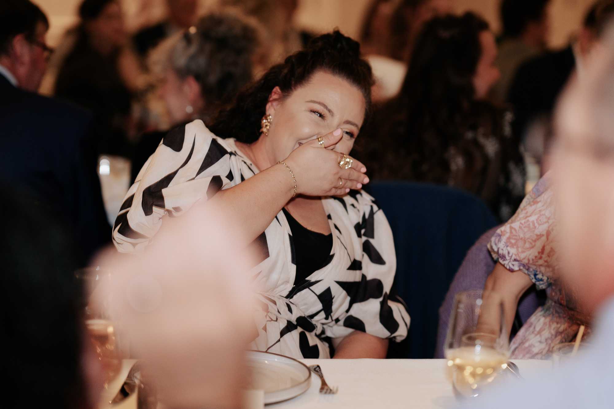 Woman with dark curly hair wearing a black and white patterned dress, laughing and covering her mouth with her hand at a formal event.