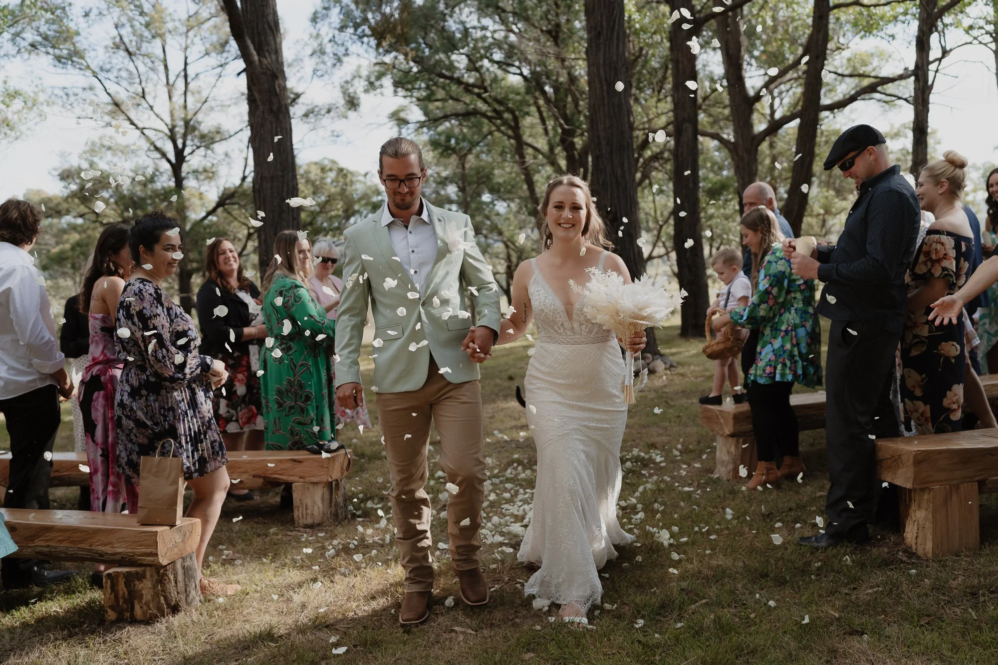 A bride and groom walking hand-in-hand down the aisle at an outdoor wedding, surrounded by guests, as flower petals are thrown in the air