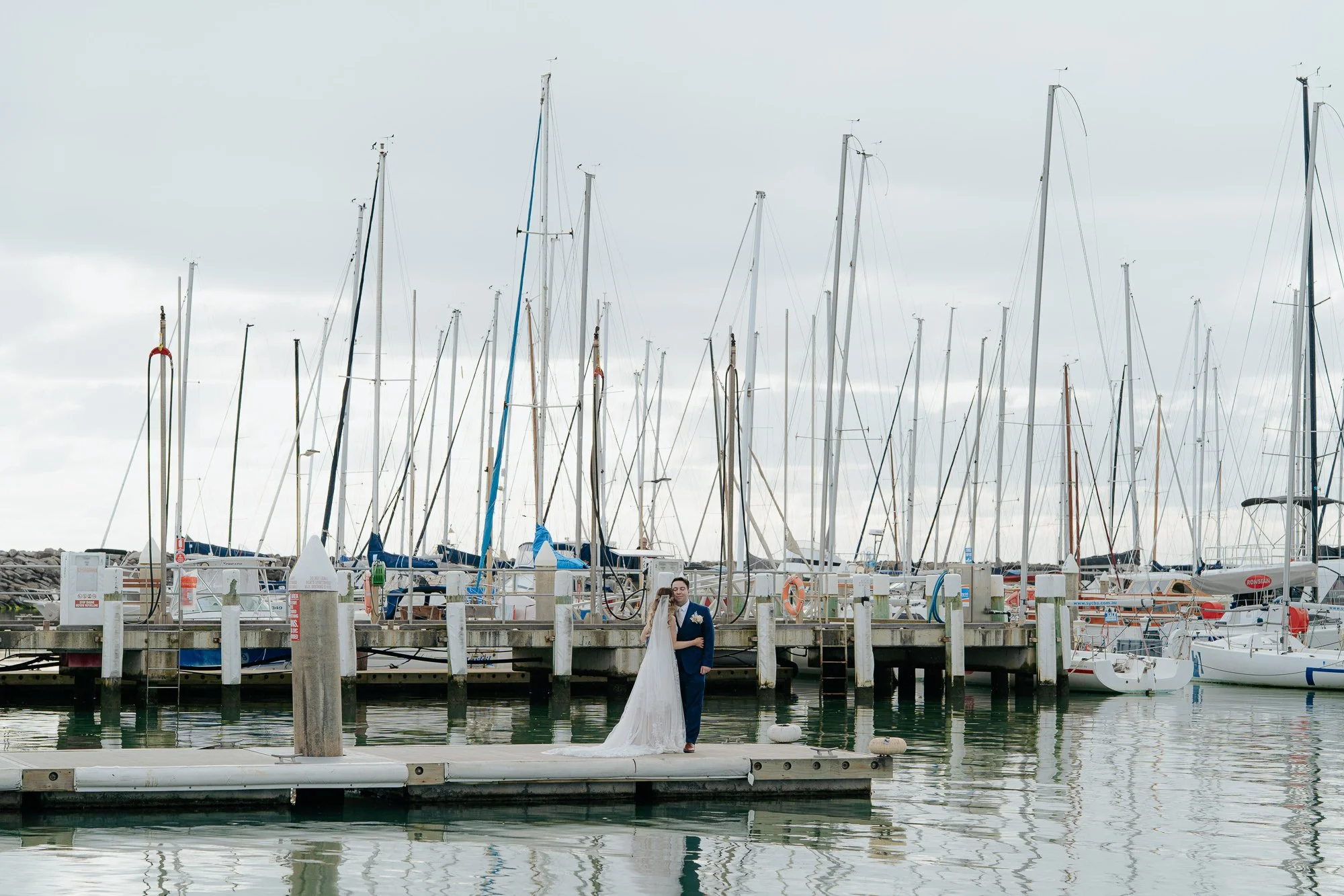 A bride and groom standing on a dock in front of sailboats at a marina on a cloudy day. Sandringham Yacht Club.