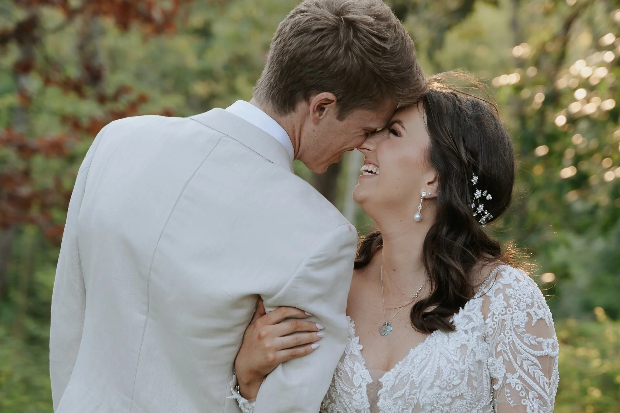 A couple dressed in wedding attire smiling and touching foreheads outdoors.
