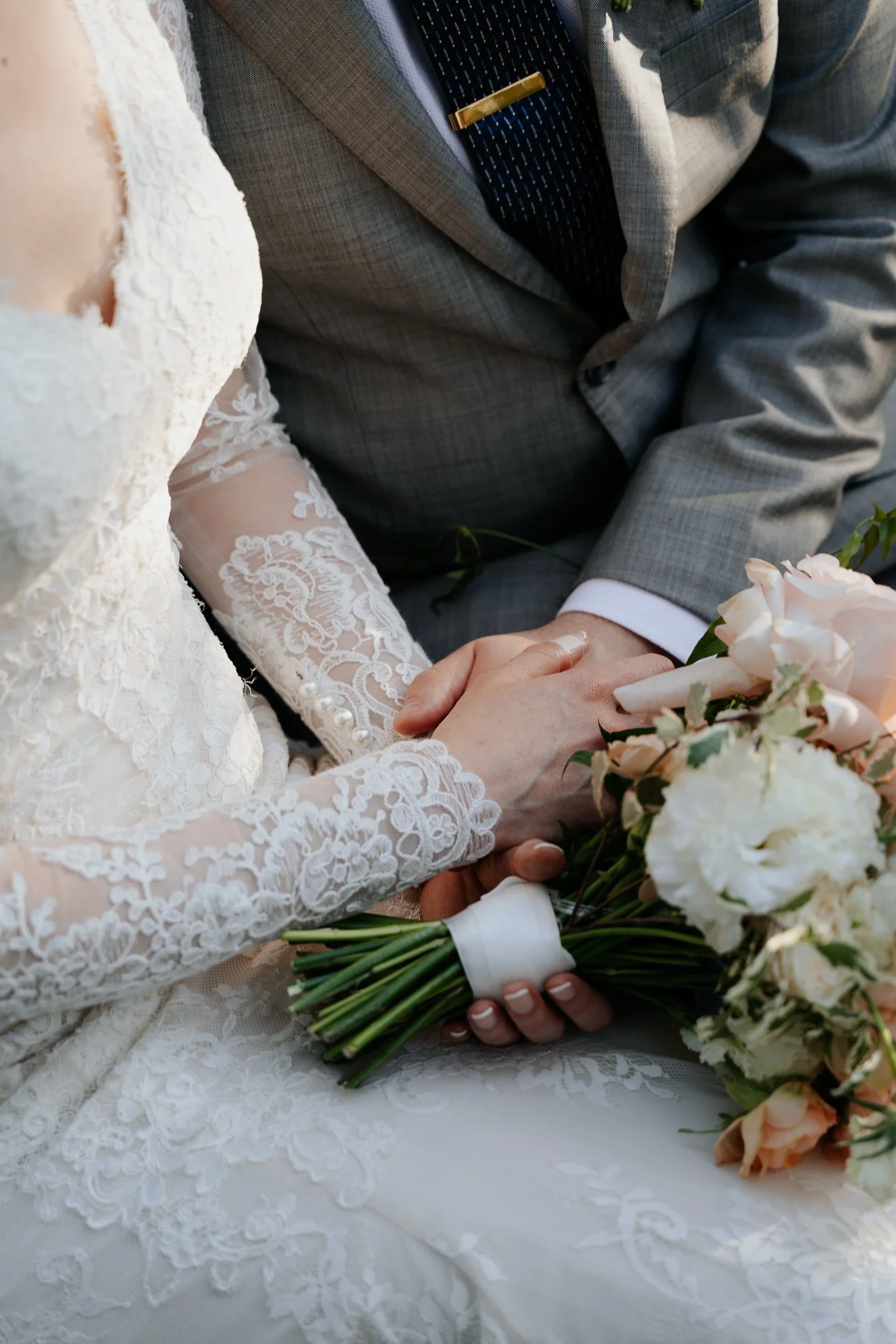 Close-up of a bride and groom holding hands, with the bride holding a bouquet of pastel pink and white roses, on their wedding day.