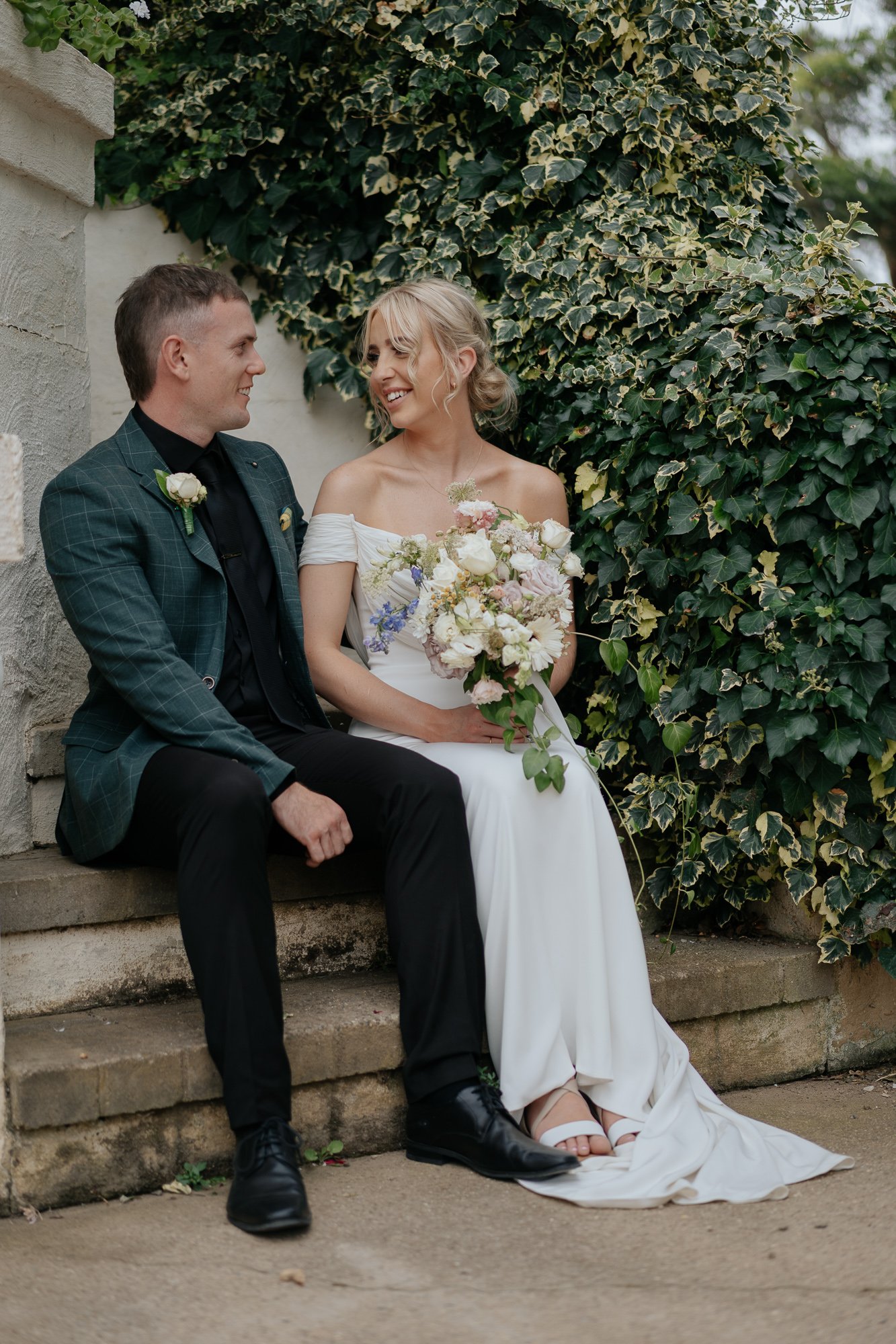 A bride and groom sitting together outdoors at Amarti at the Heritage Listed Cowwarr Butter Factory, smiling at each other, with the bride holding a bouquet of flowers, against a background of green ivy.