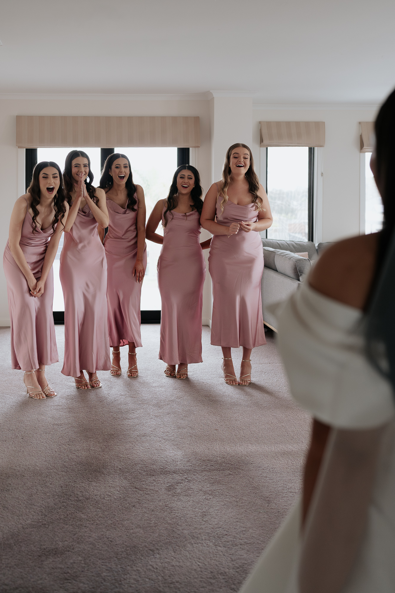 A bride sees her five bridesmaids dressed in matching pink satin dresses, reacting with joy and surprise in a room with large windows.