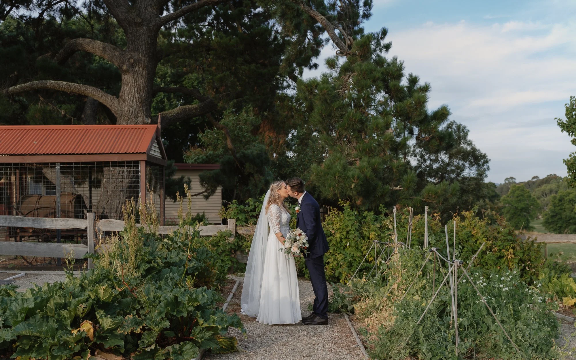 A bride and groom kiss in a garden at the Old Cheese Factory in Berwick with greenery and trees behind them, during a wedding.