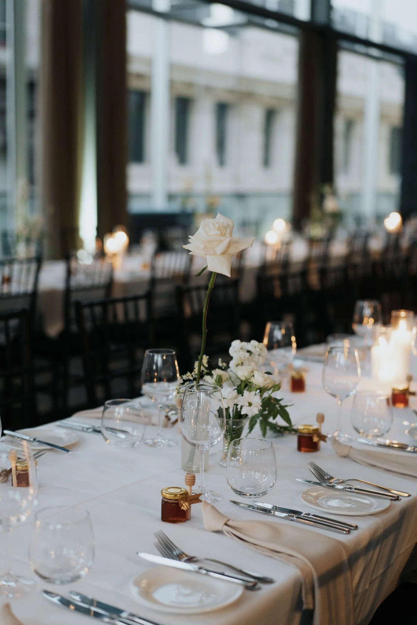 Elegant dining table with white tablecloth, glassware, silverware, small jars of honey, and a tall white rose centerpiece, set in a bright room with large windows. Alto Event Space by Damn Fine Food Co, a premium Melbourne CBD Function space.