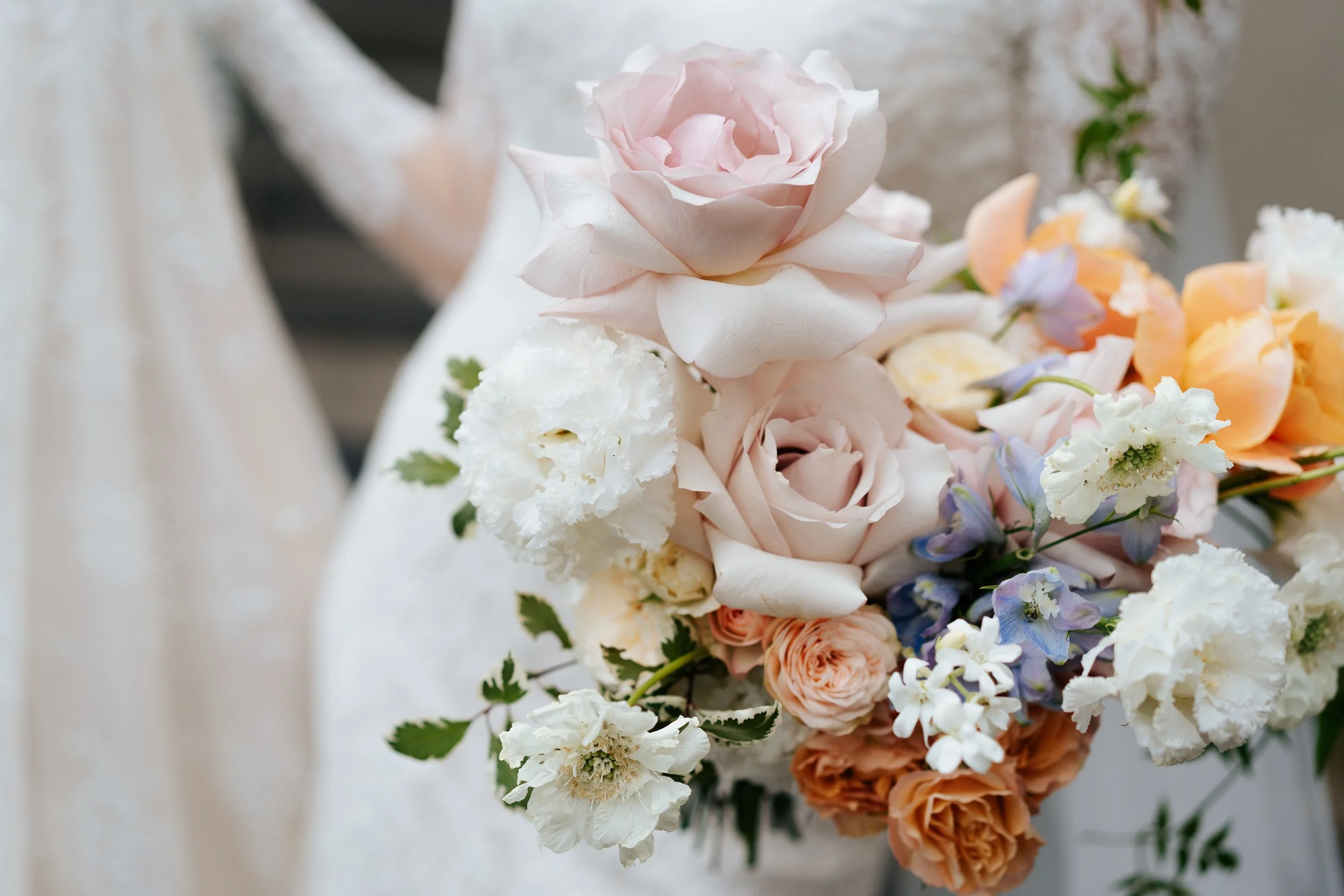 A bridal bouquet with pink and white roses, white and peach carnations, purple and white flowers, and green leaves, being held by a bride in a white lace dress.