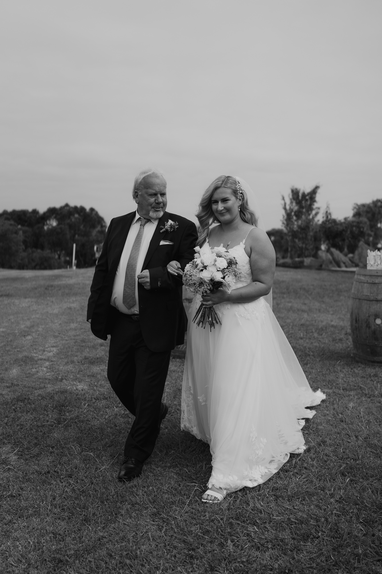 A bride in a white wedding dress holding a bouquet of flowers walking outdoors with an older man in a suit, in a grassy area with trees and barrels in the background. The Vines on Avon