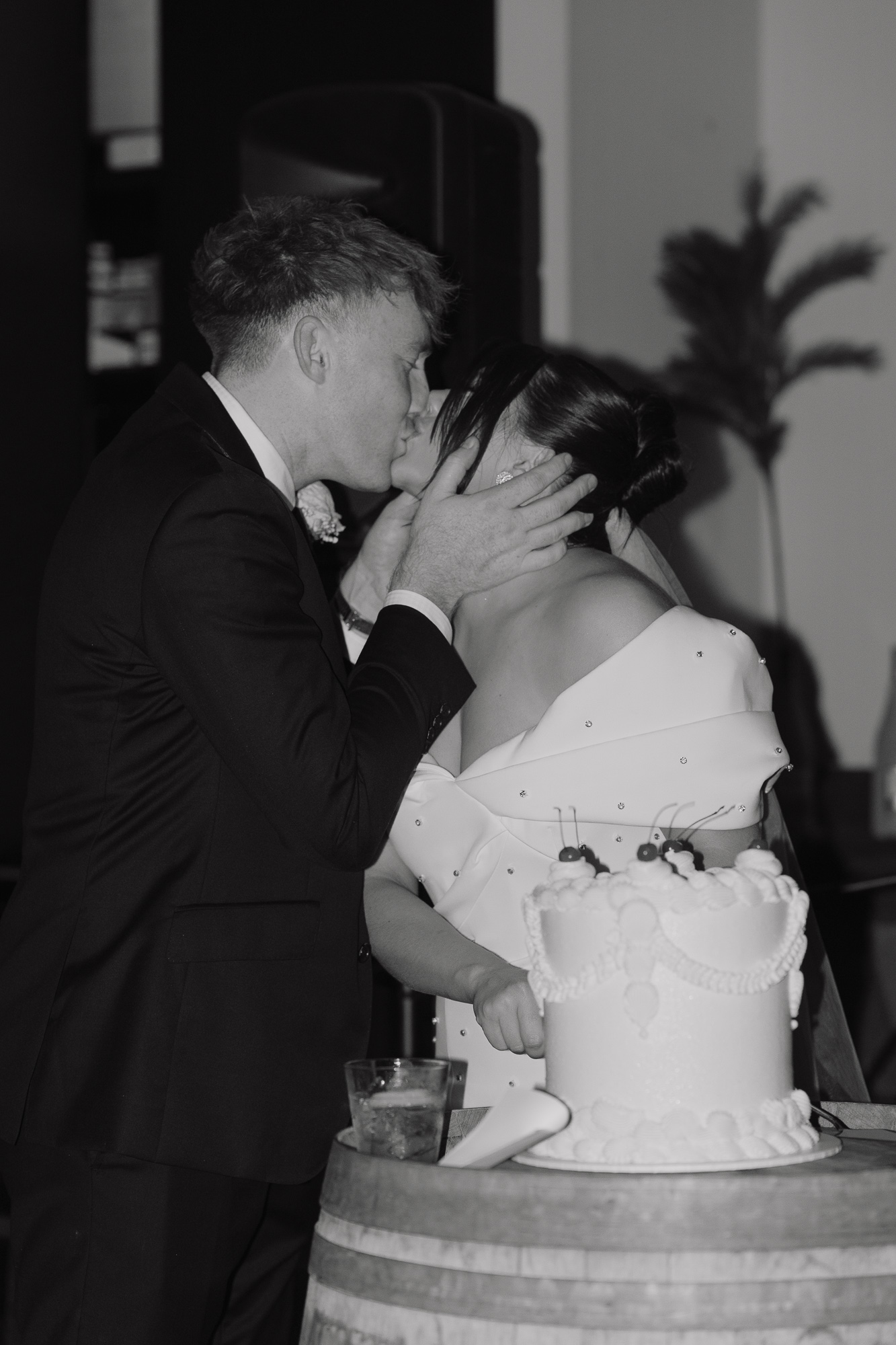 Black and white photo of a wedding couple kissing, with the groom holding the bride's face, and a wedding cake on a table in front of them.