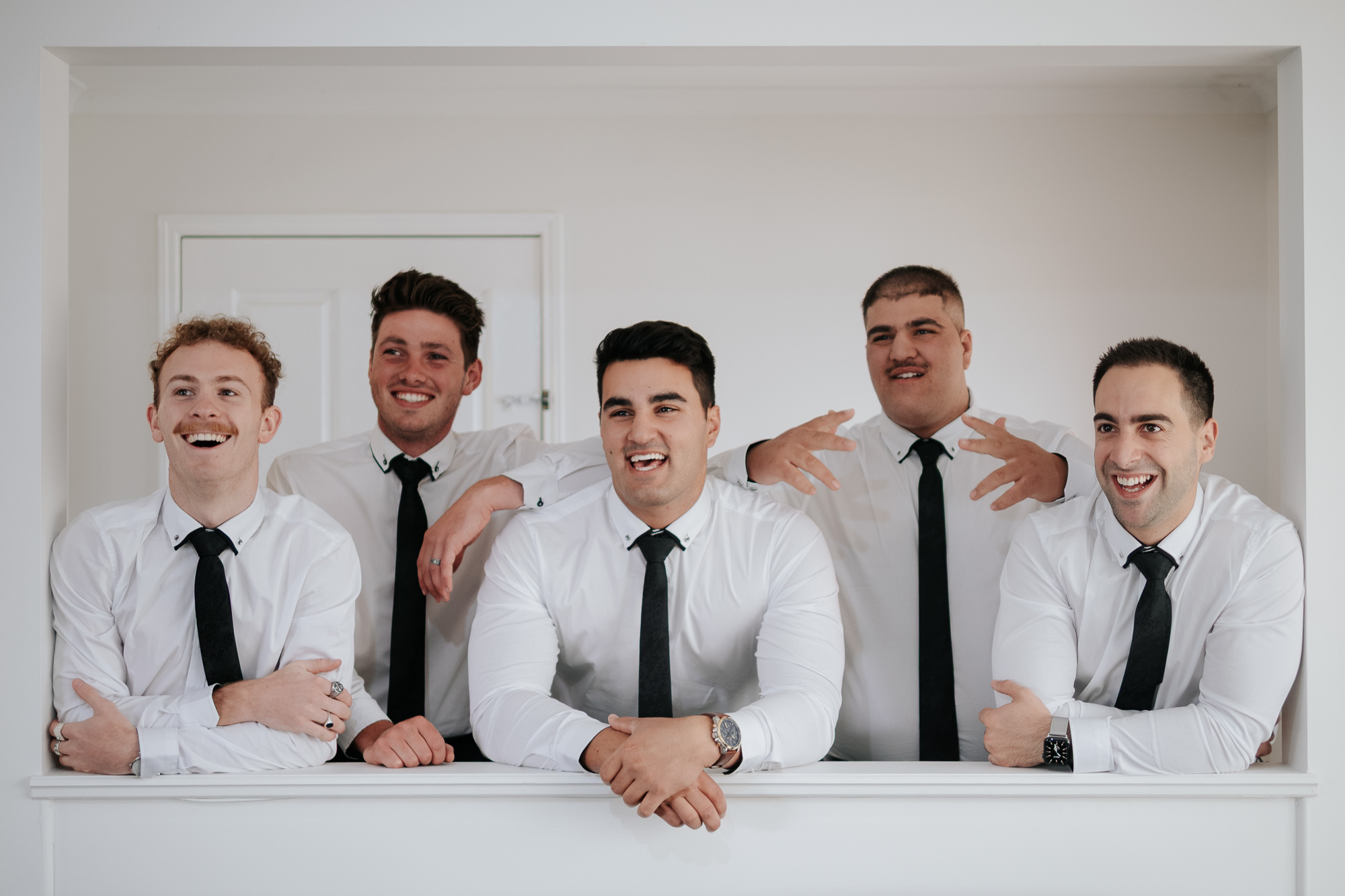 Group of five groomsmen in white shirts and black ties smiling and posing together indoors.
