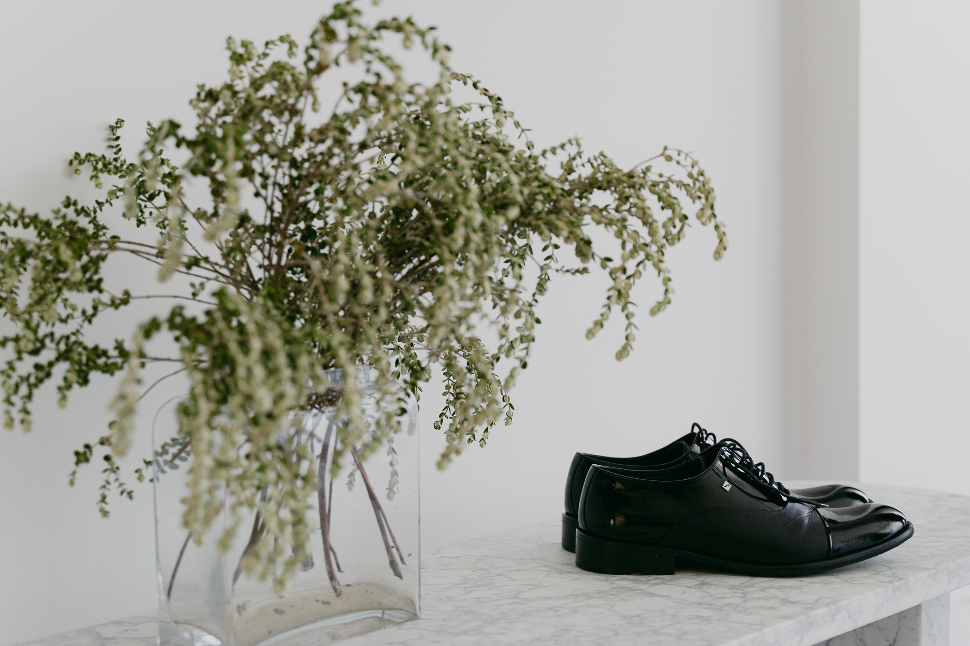 A clear glass vase with a bouquet of white and green foliage placed on a white marble table, with a pair of black leather dress shoes beside it.