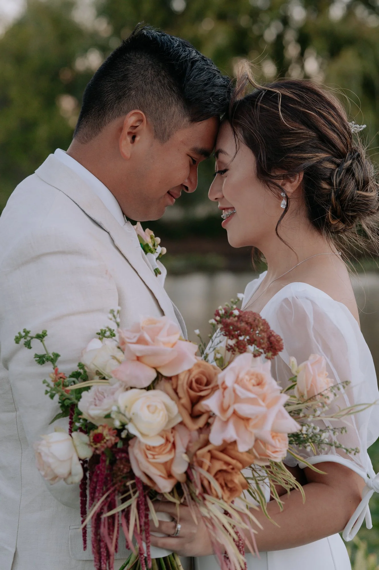 A bride and groom with foreheads touching, smiling, outdoors at Olivehouse at Greendale Grove, holding a bouquet of pink, white, and peach flowers.