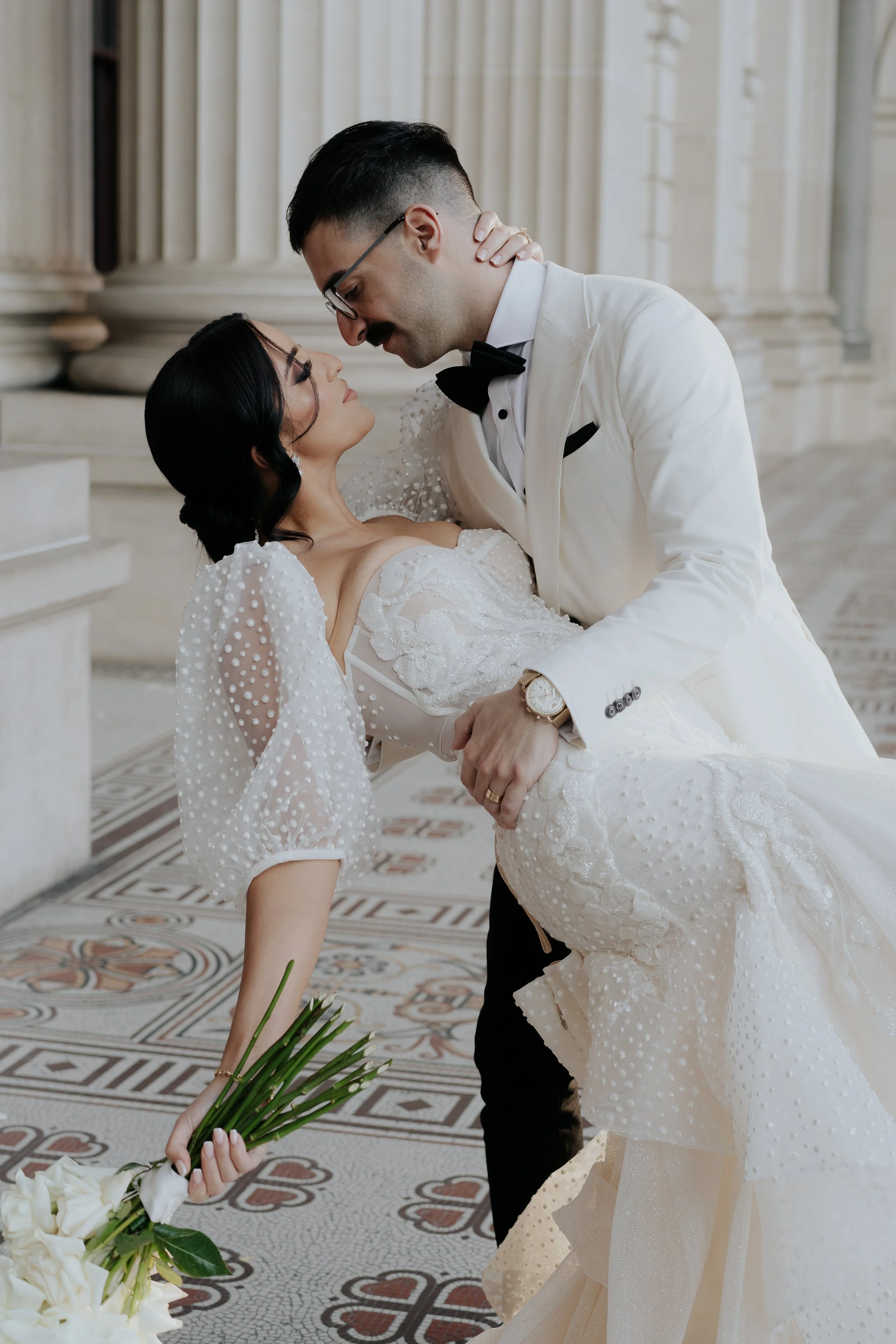A groom in a white tuxedo holding a bride in a white wedding dress in an elegant setting at Parliament House, Melbourne.