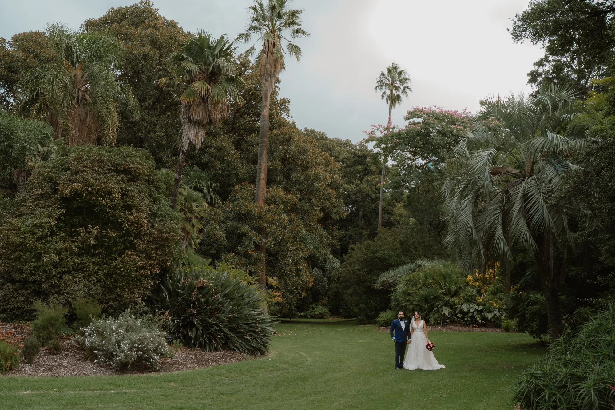 A bride and groom walking hand in hand on a lush green lawn in the Royal Botanical Gardens Melbourne surrounded by tall trees and tropical plants, with a cloudy sky above.