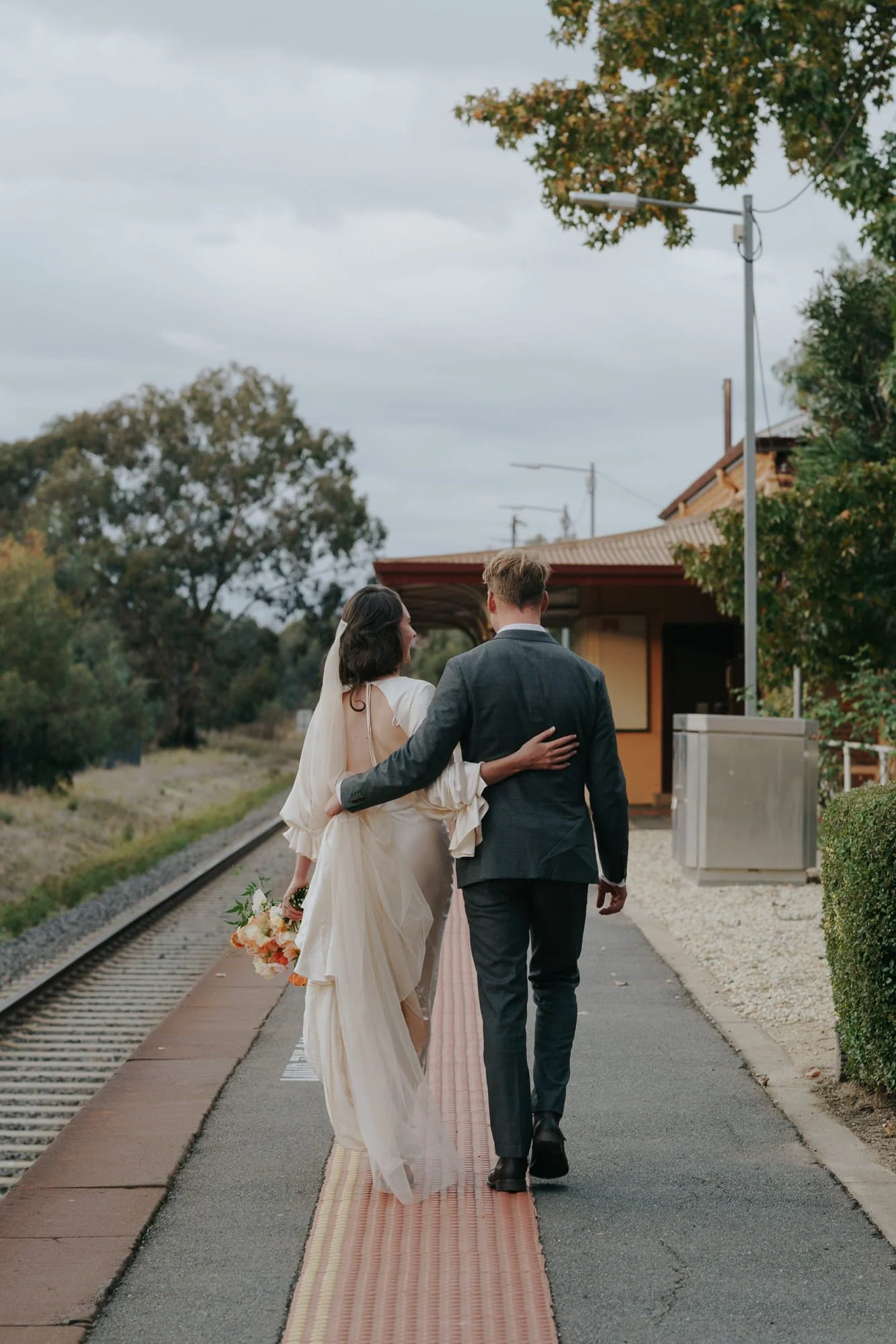 Melbourne Wedding Photography. A bride and groom walk together on a train platform, with the bride holding a bouquet of flowers and the groom wearing a suit, suggesting they are newly married.