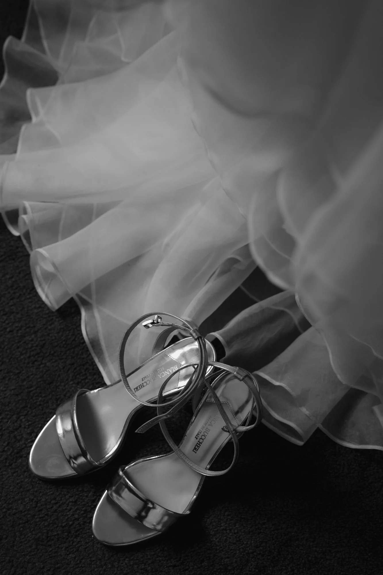 A pair of shiny high-heeled shoes, a delicate layered dress, and hoop earrings scattered on a dark textured surface, likely preparing for a special occasion or wedding.