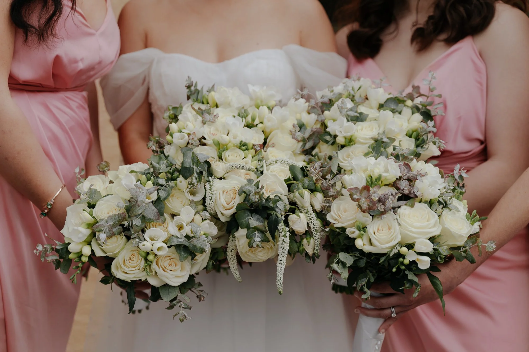 Bridal bouquet with white roses, lisianthus, greenery, and eucalyptus, held by a bride and her bridesmaids in pink dresses.