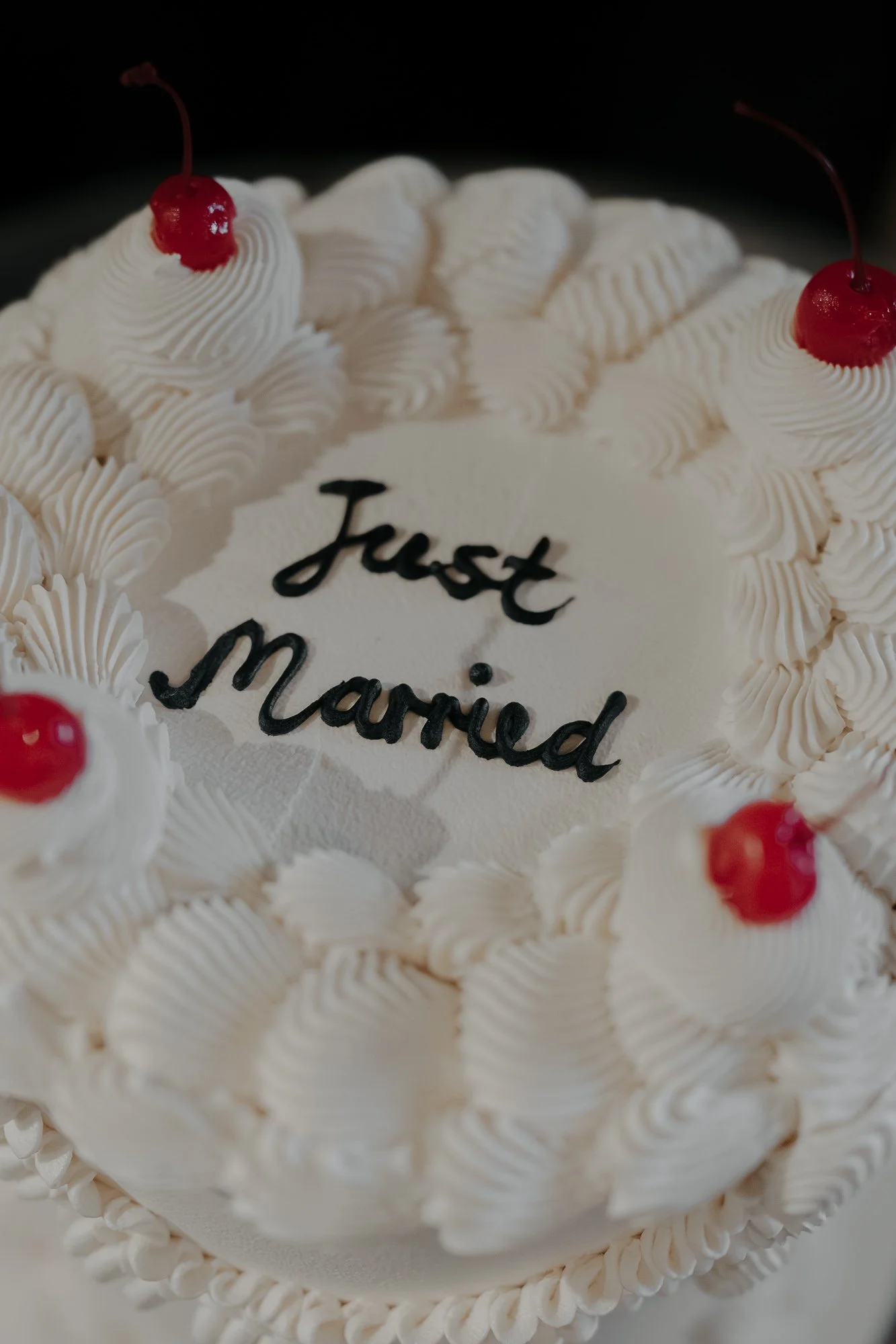 A wedding cake with white frosting and red cherries on top, decorated with piping. The cake has a message that reads 'Just Married' written in black icing.