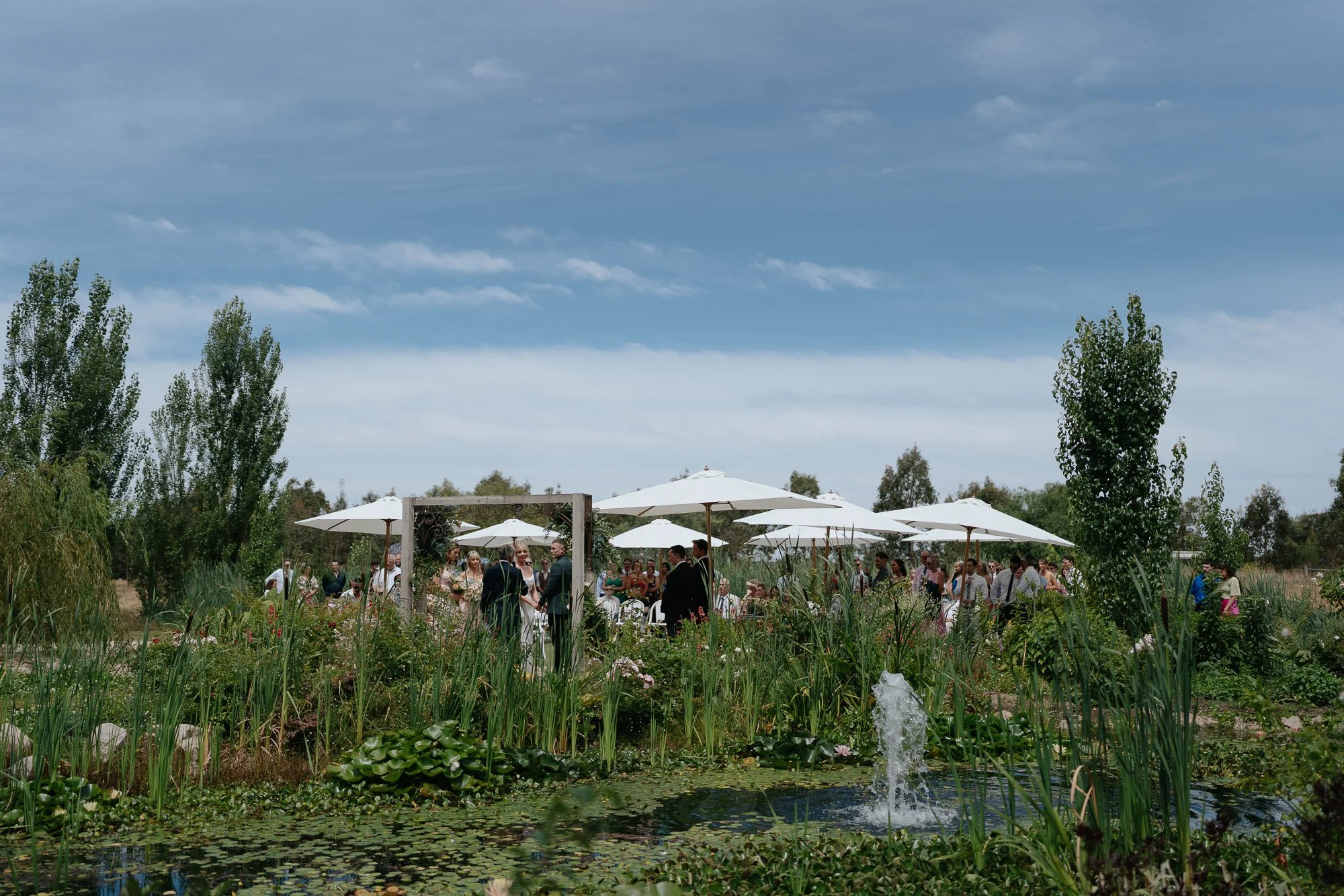 Outdoor wedding reception in a garden at Amarti at the Heritage Listed Cowwarr Butter Factory with people under white umbrellas, a pond with a fountain, tall trees, and a blue sky.