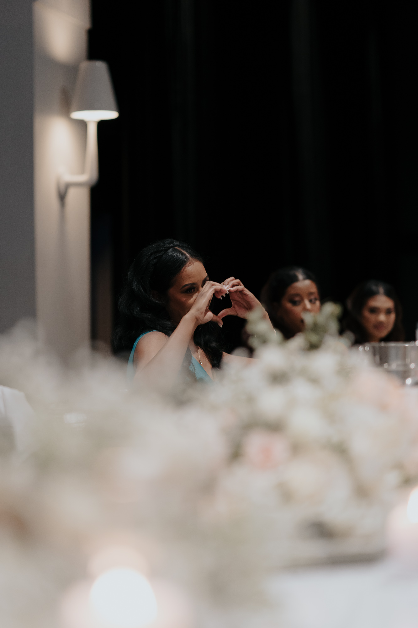 A woman at a formal event making a heart shape with her hands, seated at a table with blurred floral arrangements and other women nearby. Lakeside Events.