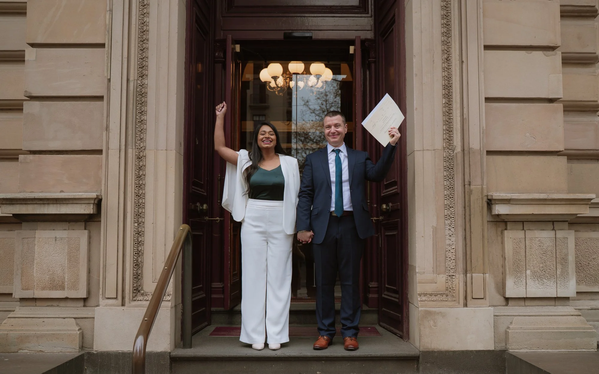 Two people, a woman and a man, standing in front of The Old Treasury Building, holding hands, celebrating, with the woman raising her arm and smiling and the man holding a document, smiling.