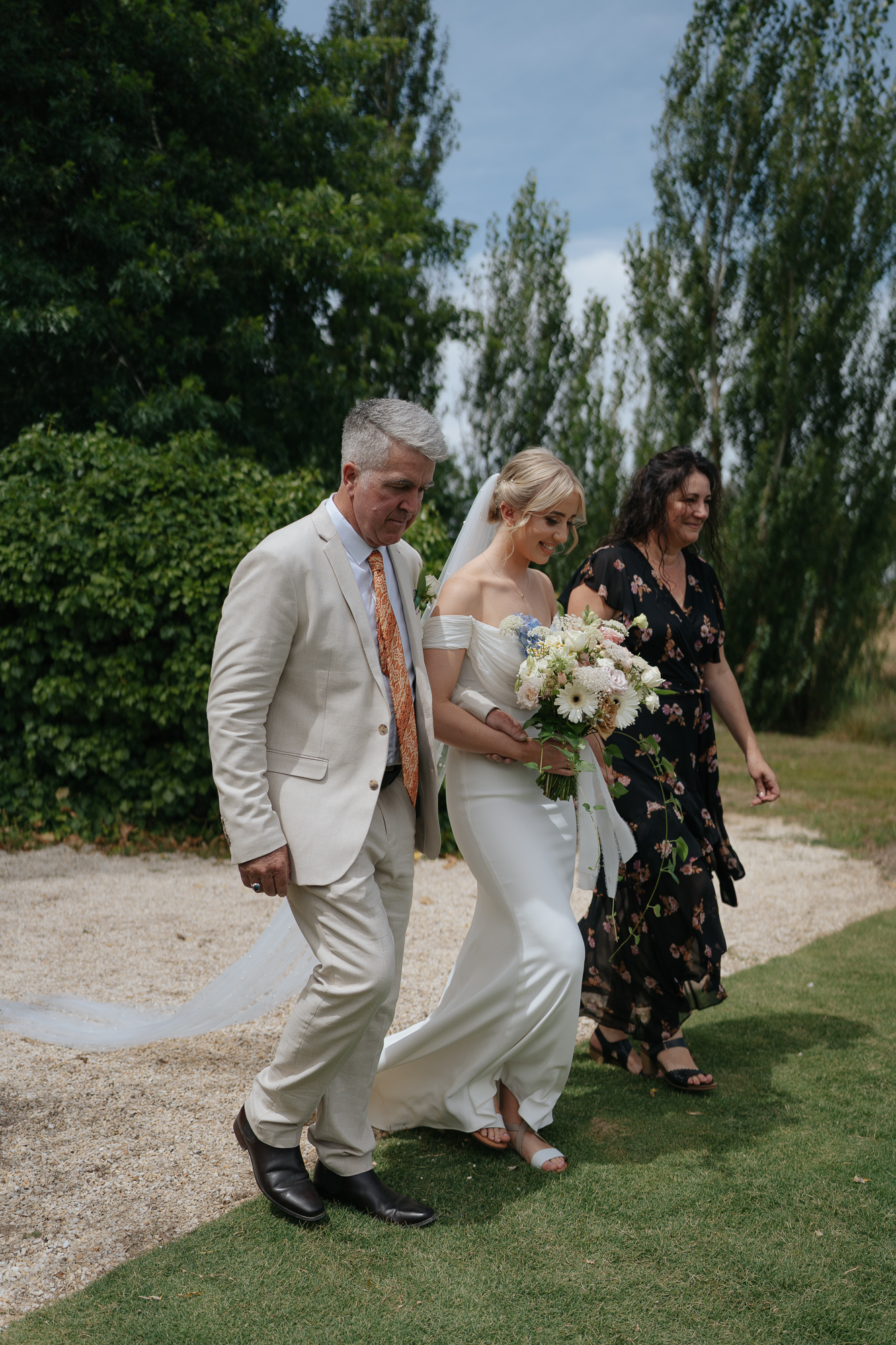 A bride in a white wedding dress holding a bouquet of flowers, walking outdoors with an older man in a light suit and a woman in a black floral dress, surrounded by greenery.