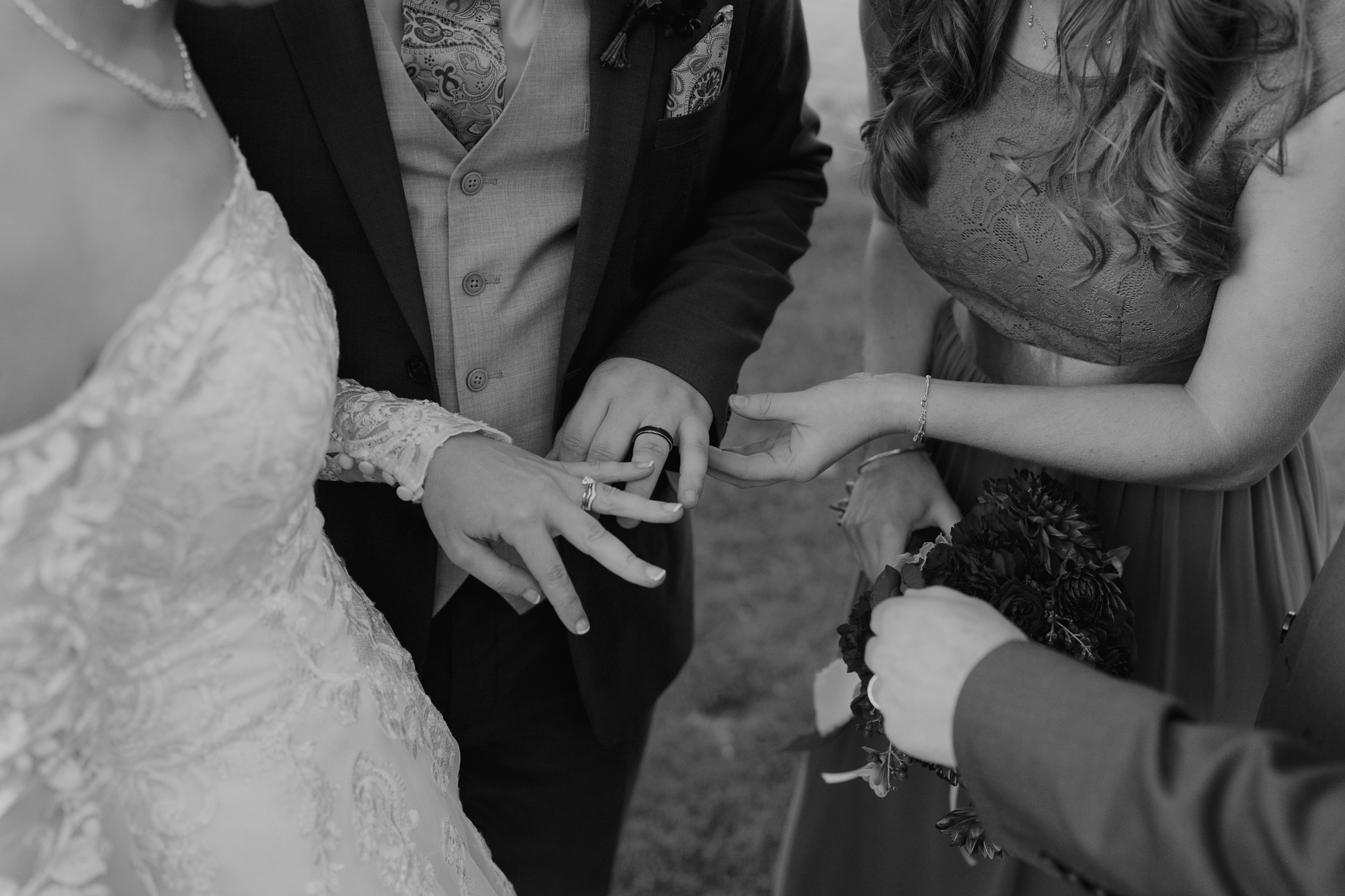 Black and white photo of a wedding ceremony showing a bride and groom exchanging rings with a bridesmaid and groomsman assisting, focusing on their hands.