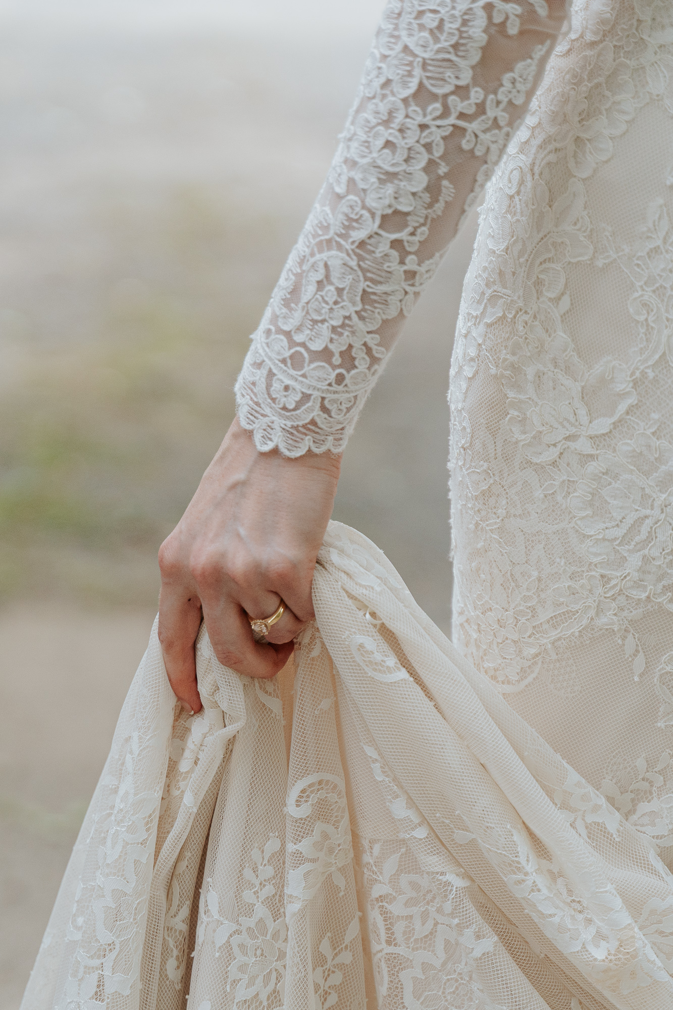 Close-up of a bride's hand holding the lace hem of her wedding dress, showing intricate lace details on the dress and a wedding ring. Chateau Wyuna