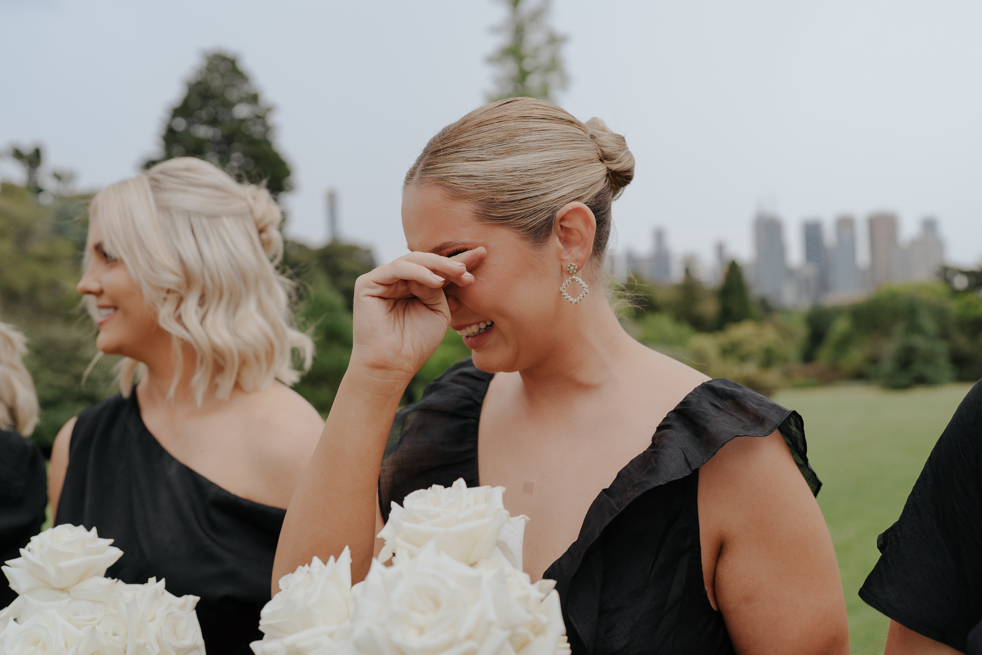 A woman in a black dress smiling and covering her face with her hand, standing outdoors with greenery and city skyline in the background, holding a bouquet of white roses. Royal Botanic Gardens Melbourne