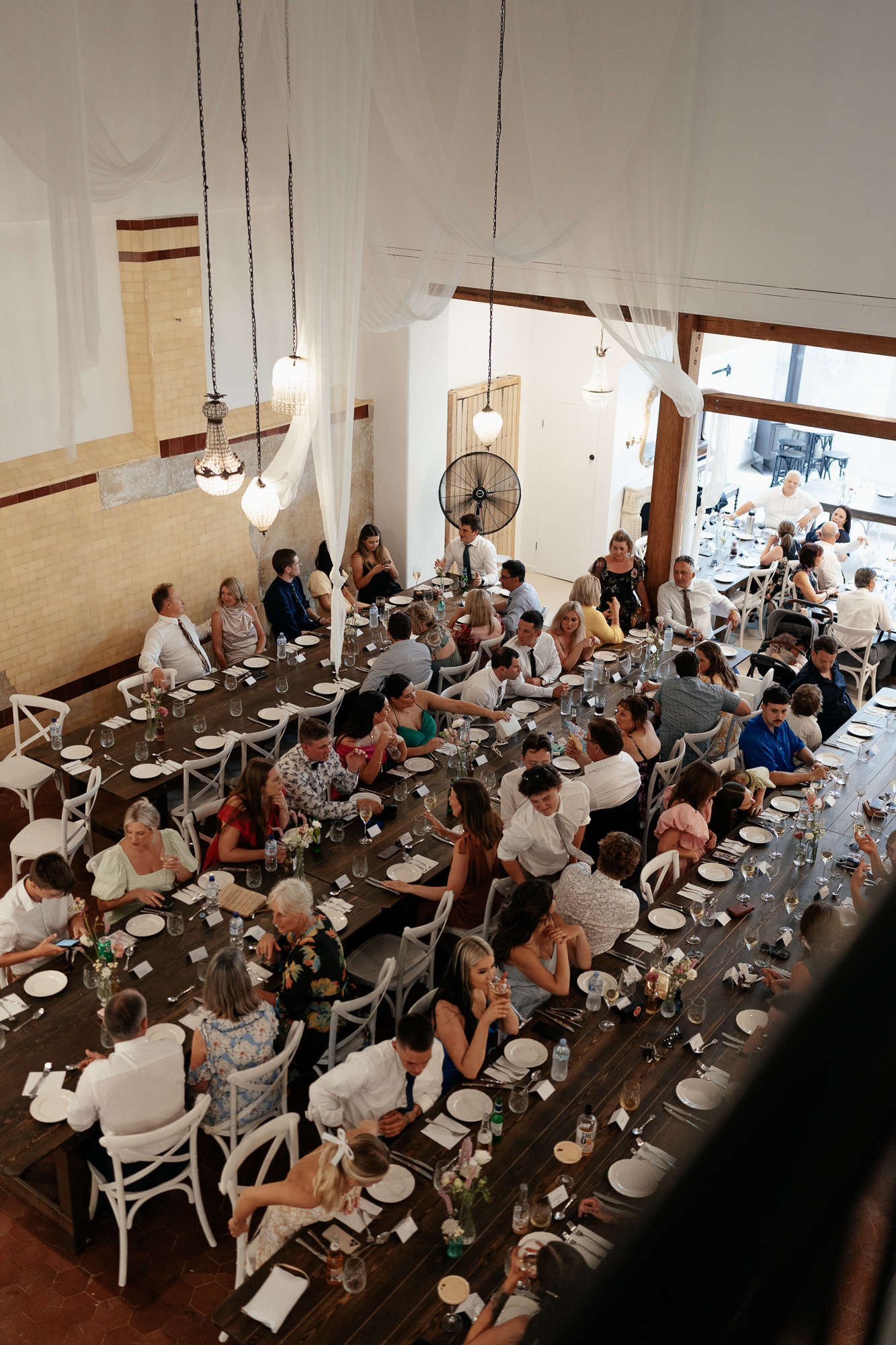 A large gathering of people sitting at long dining tables set for a formal event in a spacious, decorated room with hanging lights and draped curtains. Amarti Wedding Venue historic Cowwarr. 