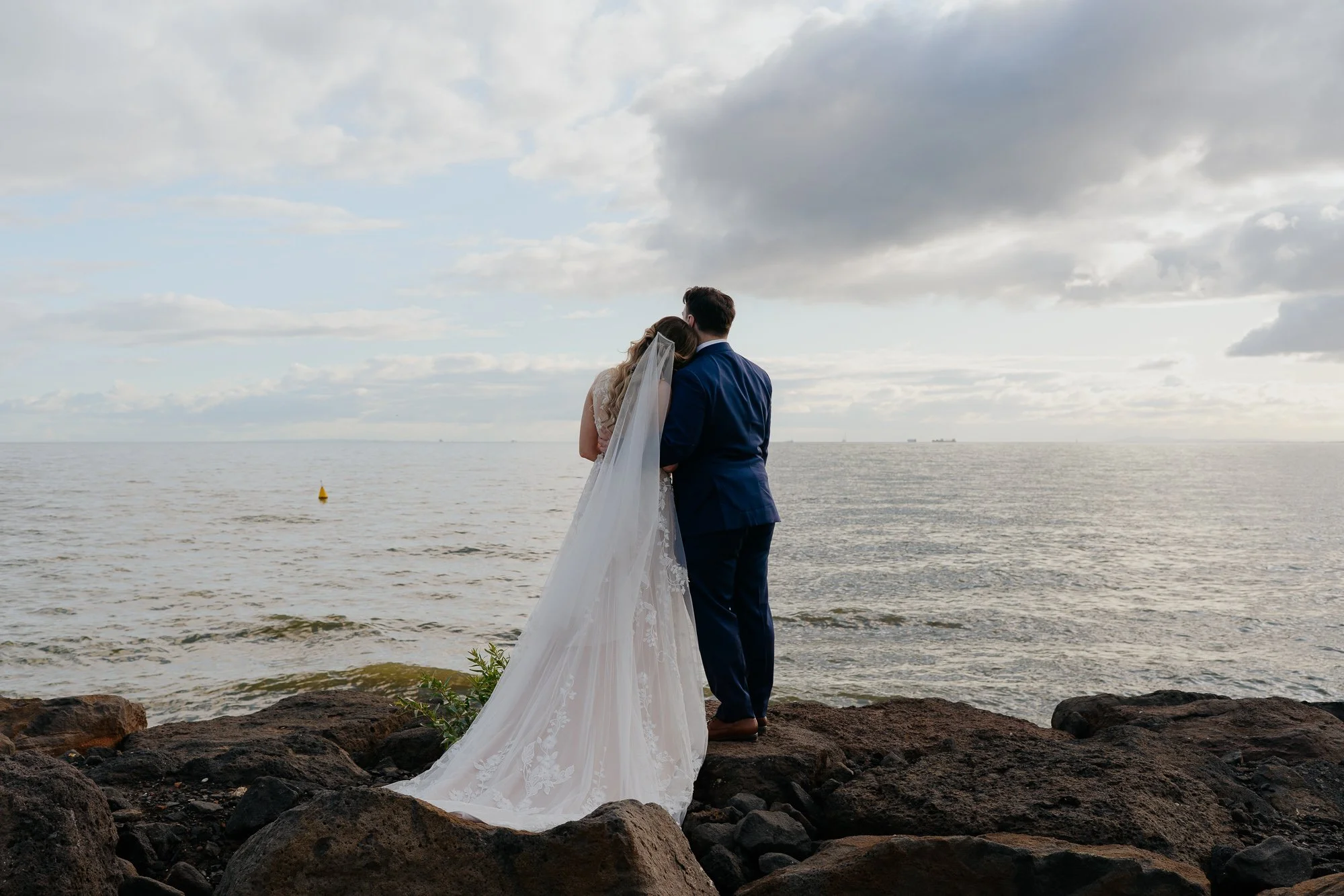 Bride and groom holding each other on rocks by the water, facing the ocean during sunset at Sandringham Yacht Club.