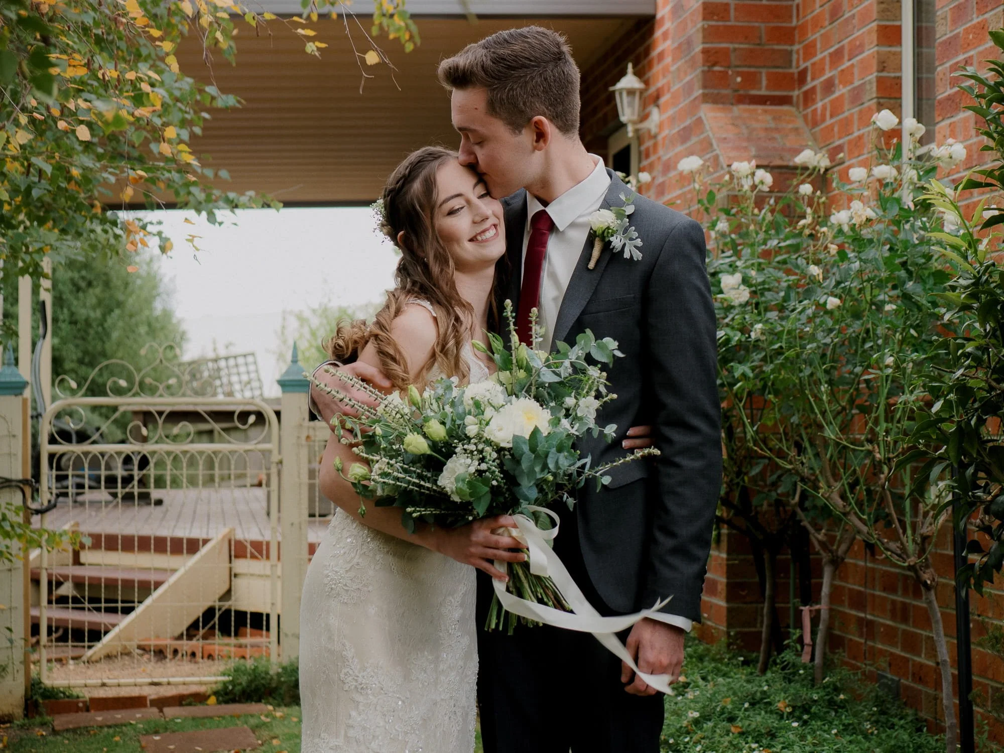 Melbourne Elopement Photography. A bride and groom standing outdoors in front of a house, embracing and smiling. The bride is holding a bouquet of white and green flowers, and the groom is wearing a black suit with a boutonniere. The bride has long, 