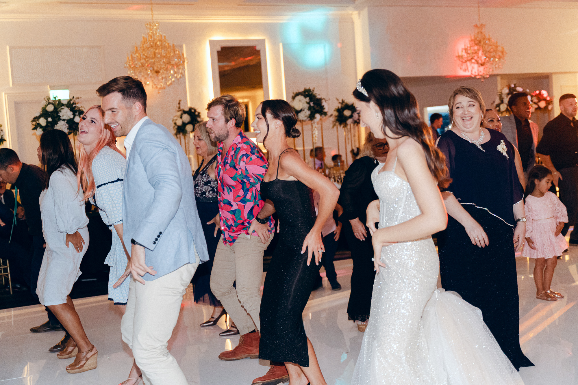 People dancing at a wedding reception in an elegant decorated ballroom with chandeliers and floral arrangements.