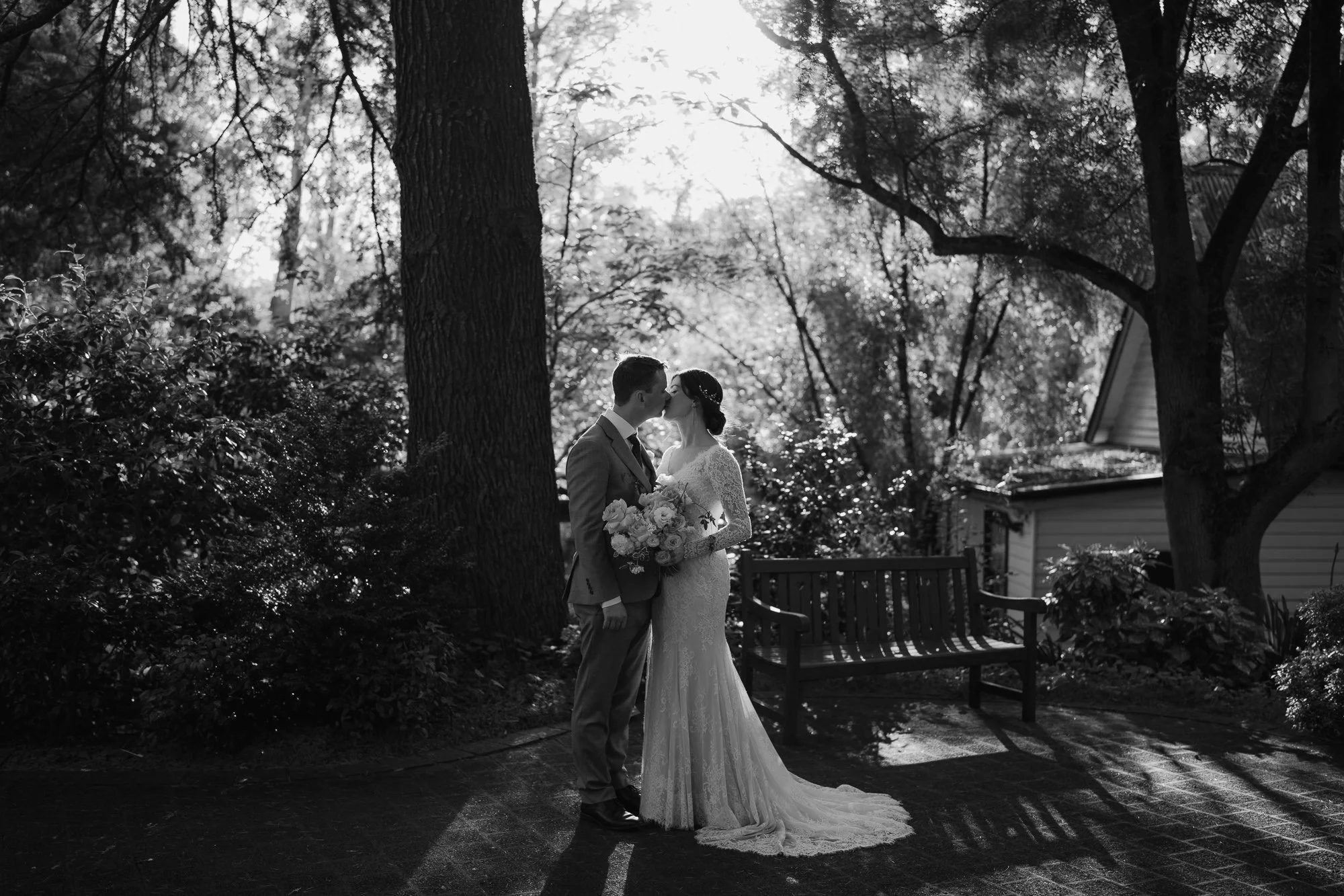 Black and white photograph of a bride and groom sharing a kiss in a garden at Chateau Wyuna with trees, bushes, and a wooden bench in the background.