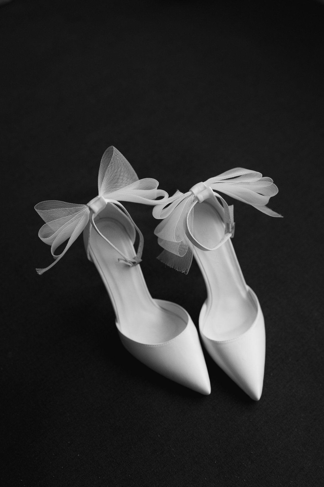 Black and white photograph of a pair of white high heels with pointed toes and decorative bows made of tulle on the ankle straps, against a black background.