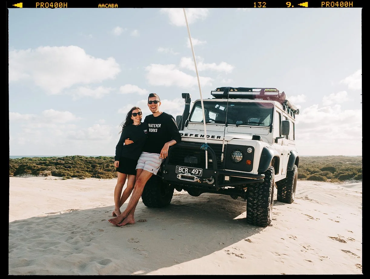 A man and woman standing next to a white off-road vehicle on a sandy beach with a clear sky and scattered clouds.