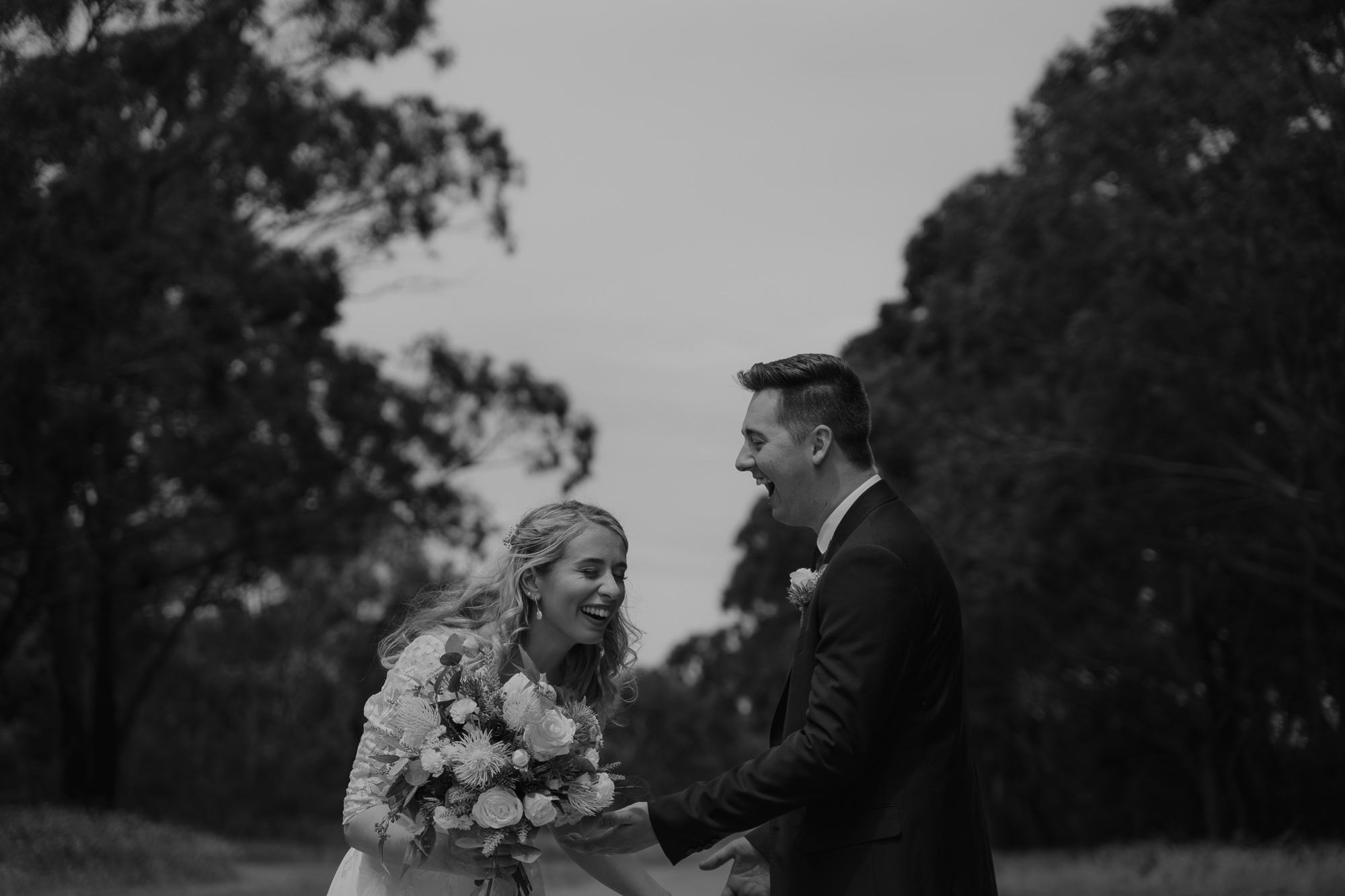 A black-and-white photo of a bride and groom sharing a joyful moment outdoors, with trees in the background. The bride holds a bouquet of flowers and laughs, while the groom, in a suit, smiles and looks at her.