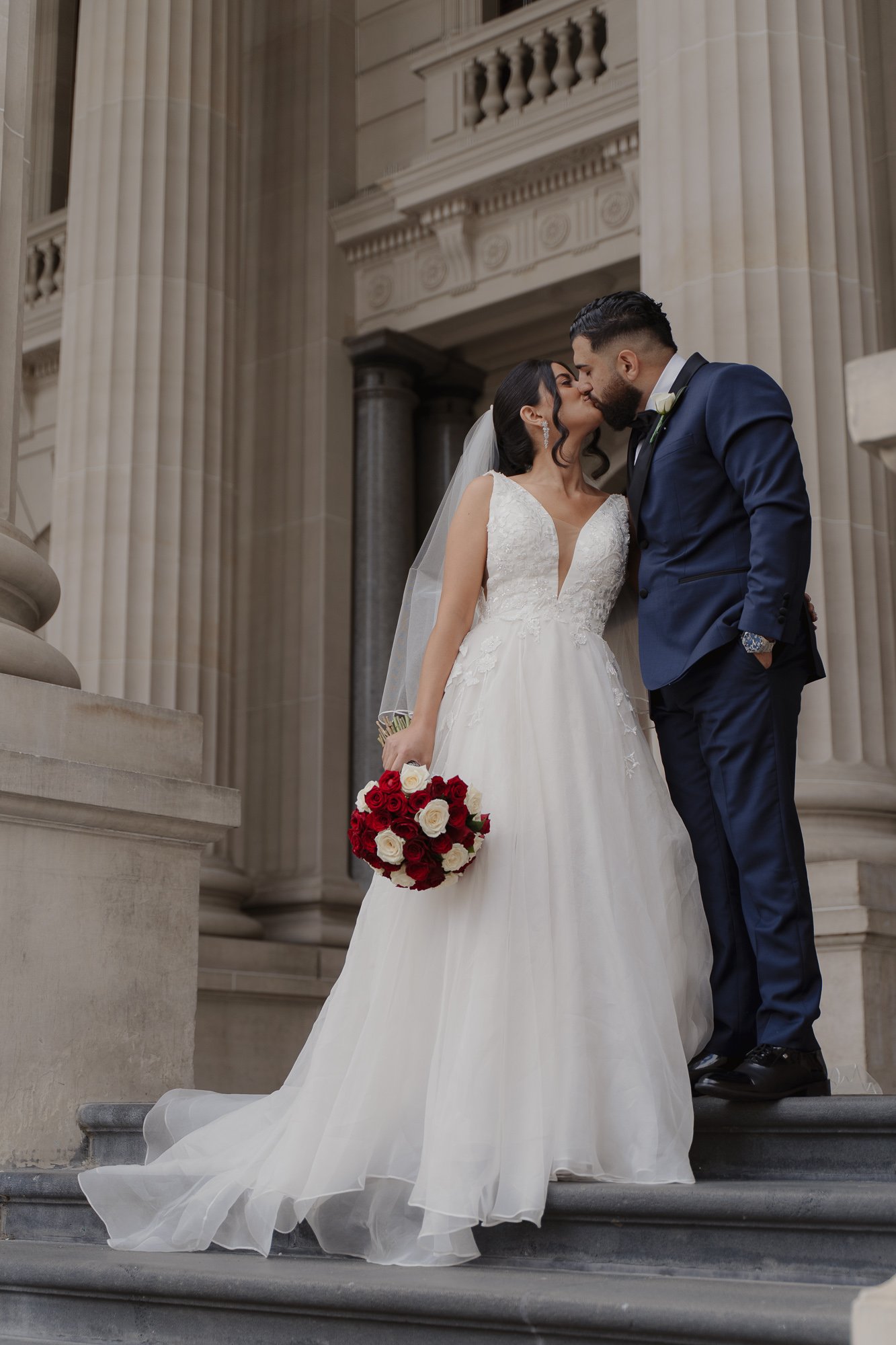 A bride and groom kissing on steps of a neoclassical building with large columns, the bride holding a bouquet of red and white roses.