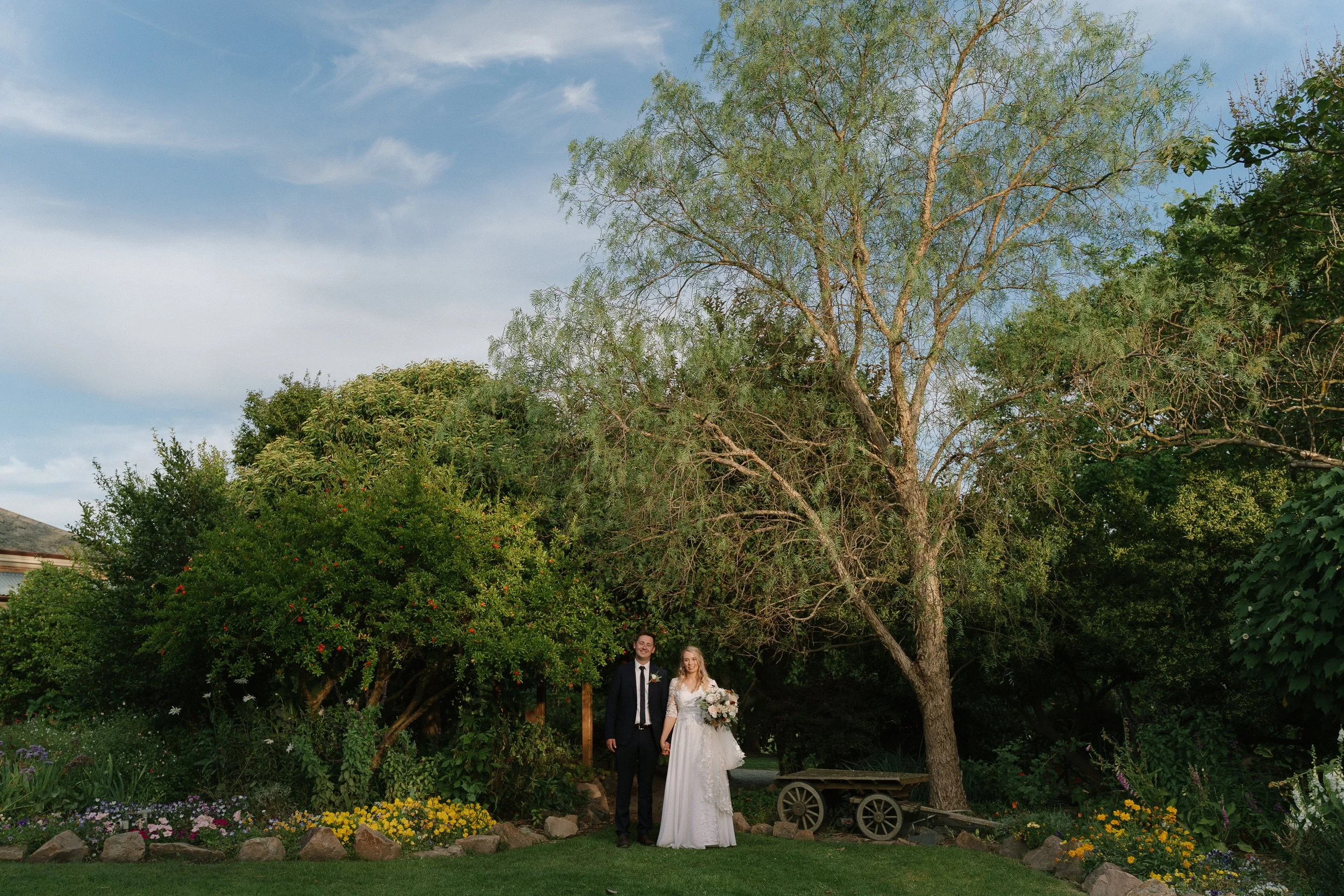 A bride and groom standing hand in hand outdoors in a garden, with trees and flowers around them, under a partly cloudy sky at the Old Cheese Factory, Berwick.