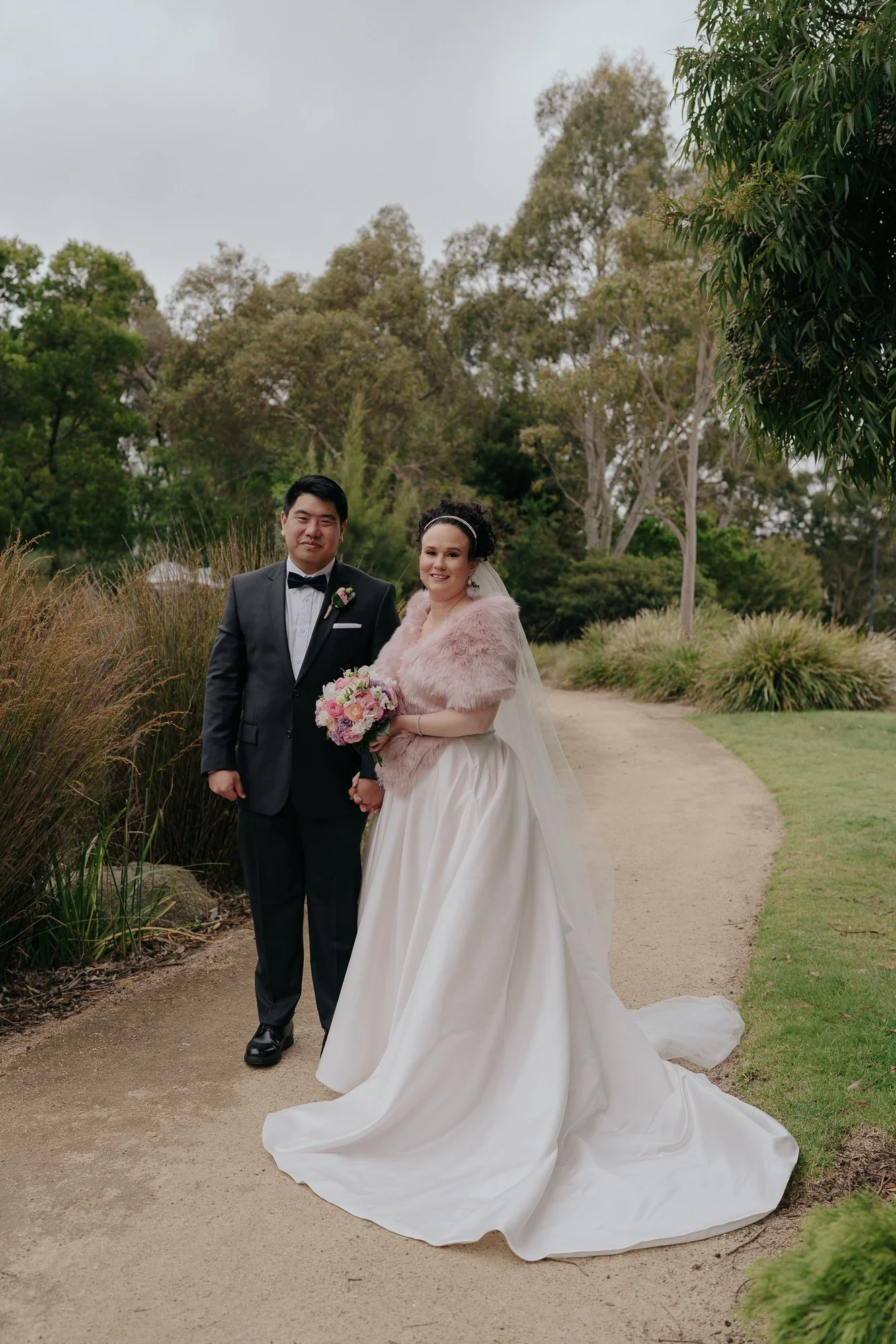 A bride and groom standing together outdoors on a garden path; the bride is wearing a white wedding dress with a long train and a pink fur shawl, holding a bouquet of pink and purple flowers, while the groom is dressed in a black tuxedo with a bow ti