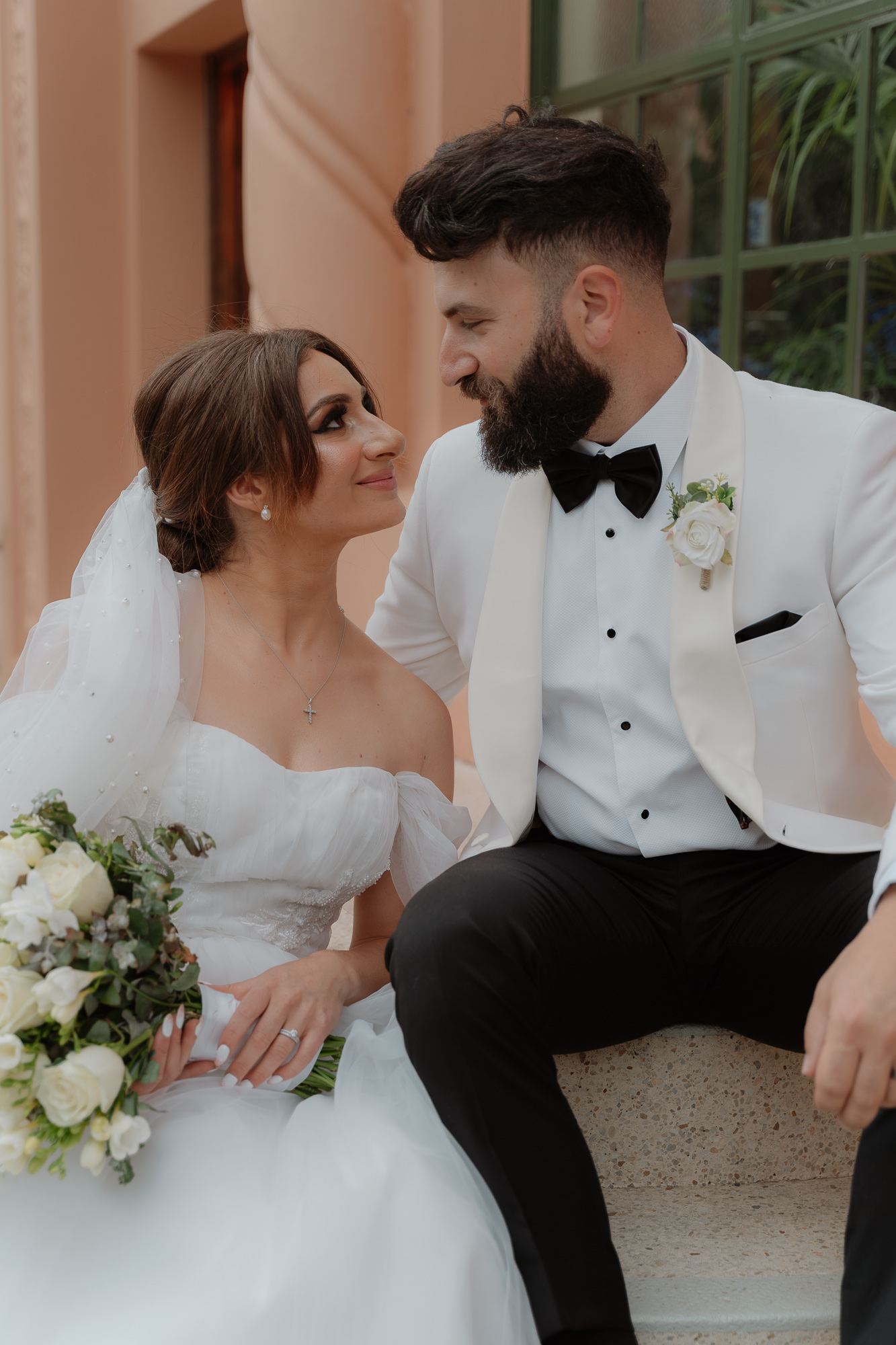 A bride and groom sit close together, gazing into each other's eyes during their wedding, with the bride holding a bouquet of white roses and greenery, and the groom wearing a white tuxedo with a black bow tie and boutonniere.